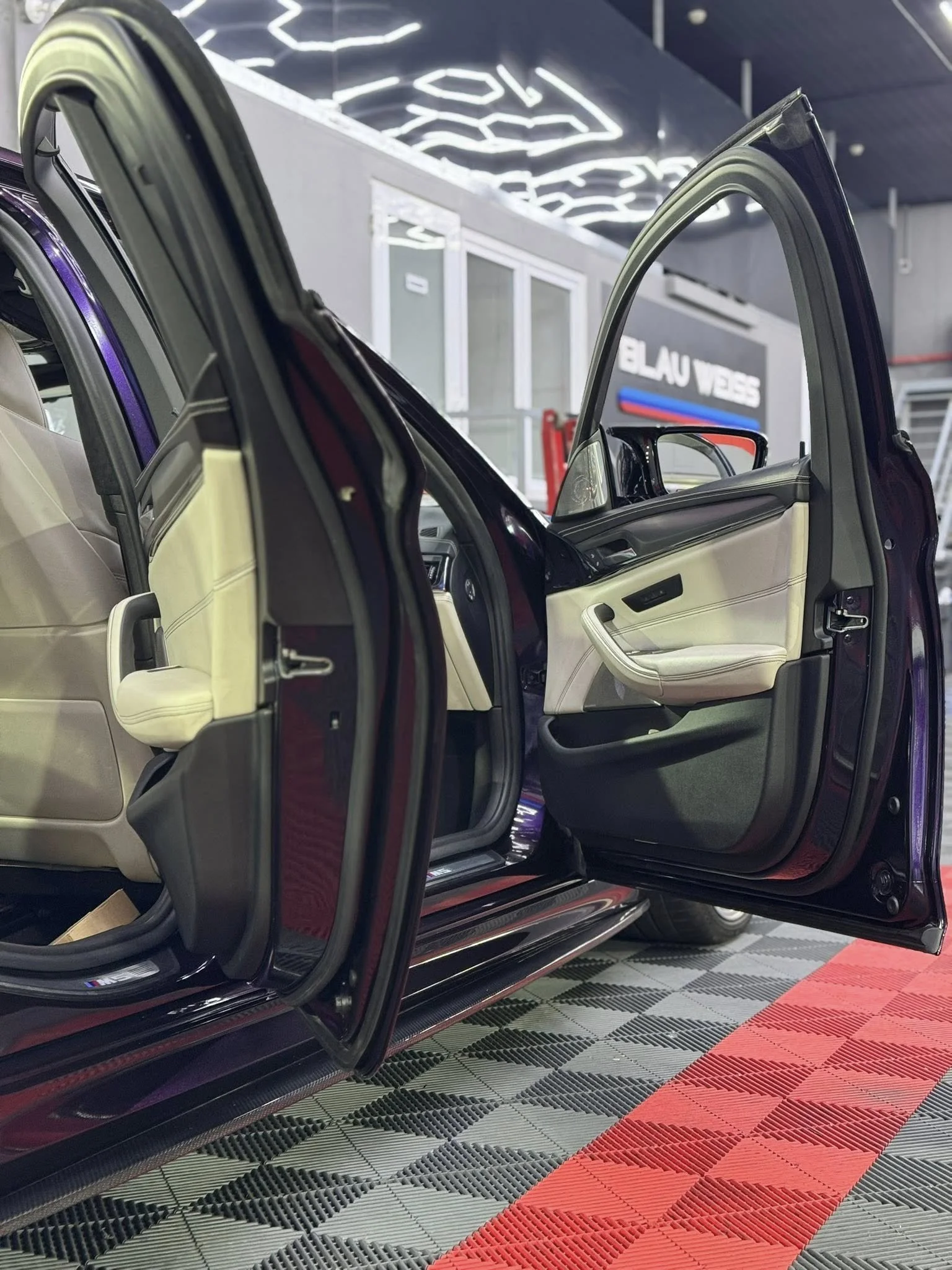 Open car doors showing the beige interior of a black vehicle inside a showroom with modern lighting and a sign reading 'BLAU VELLES' in the background.