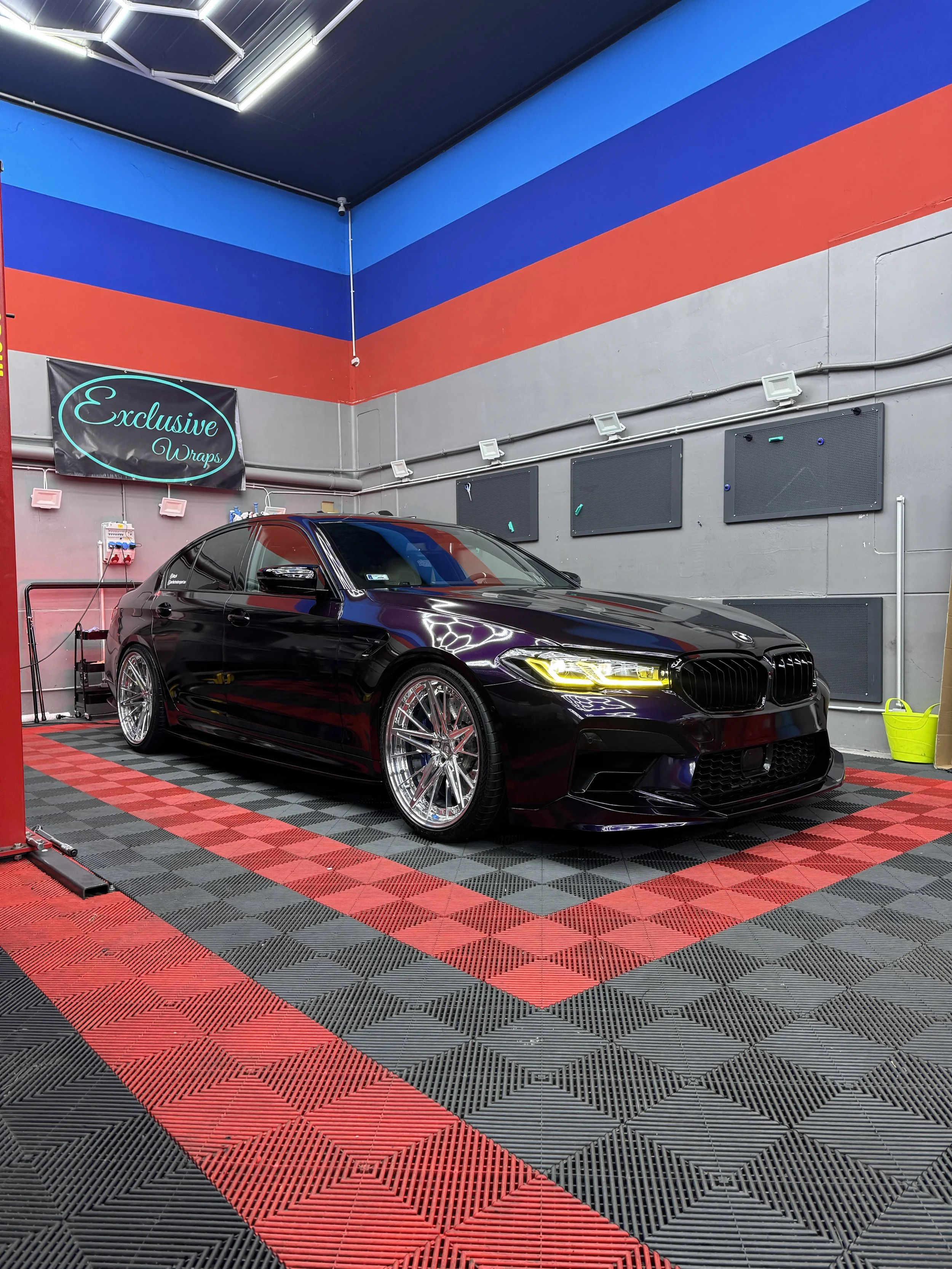 A black BMW sedan with tinted windows and shiny chrome wheels parked inside a colorful car detailing shop. The shop has a black, red, and blue striped wall, with a neon sign reading 'Exclusive Wraps' and several black panels on the wall.