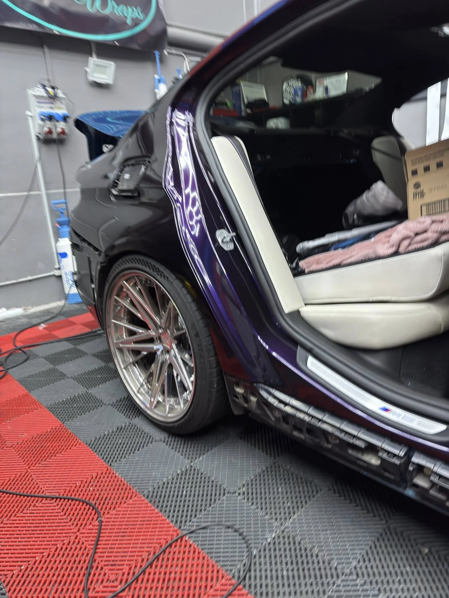 Close-up of the rear side of a shiny black sports car with aftermarket wheels, open rear door, and a view of the interior and garage background.