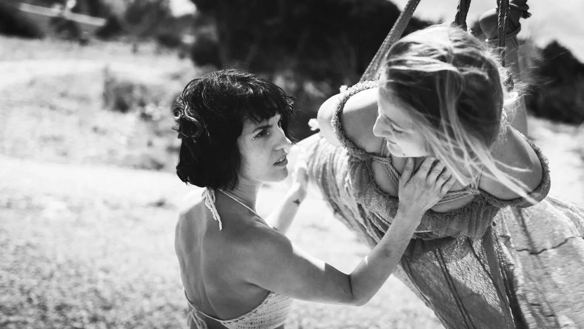Two women with short and long hair engage in an intense stare, with one holding the other's shoulder, outdoors on a sunny day.
