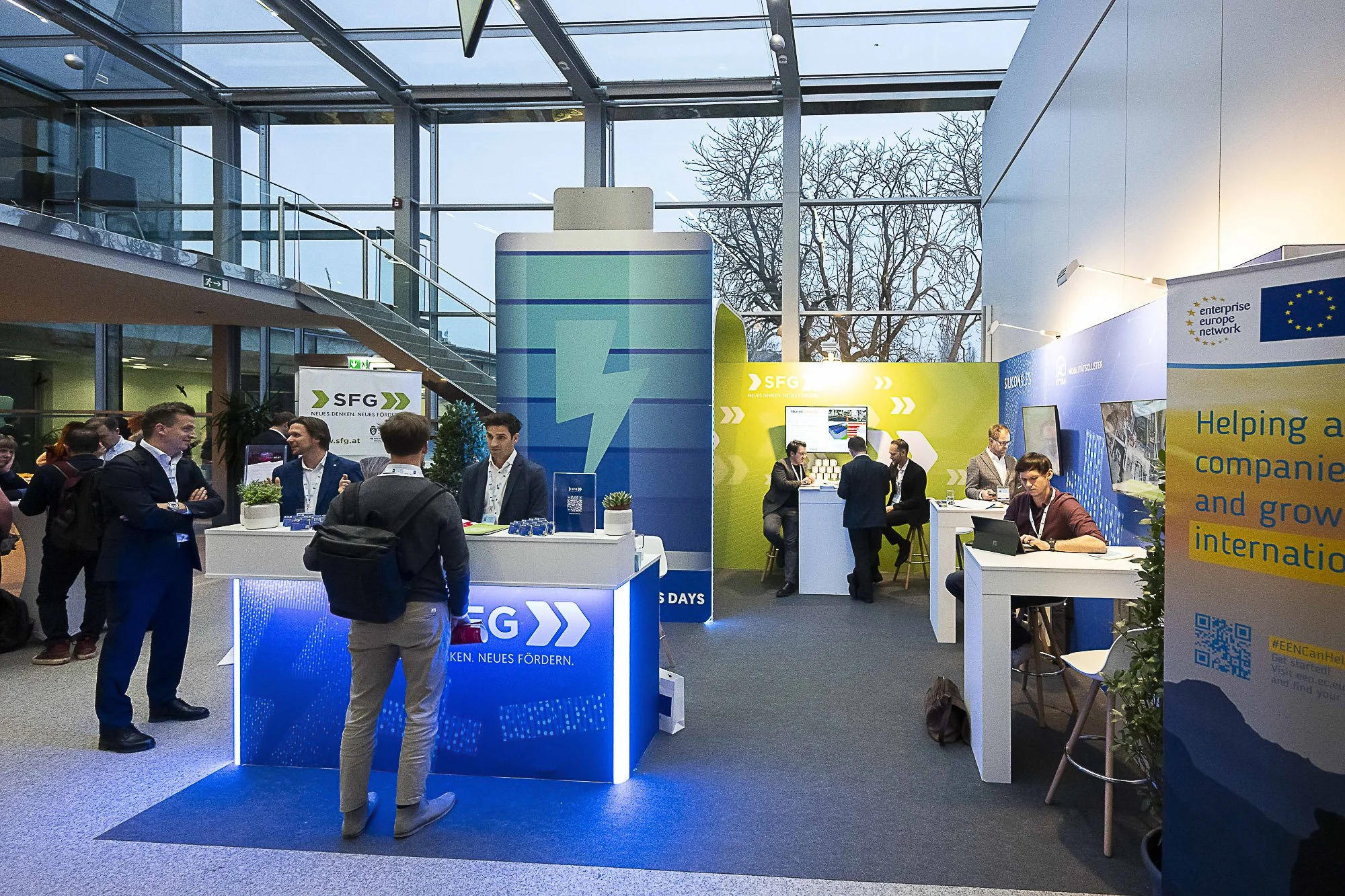 Attendees at a business conference booth display, with banners and informational materials, inside a modern convention center with glass walls and a high ceiling.