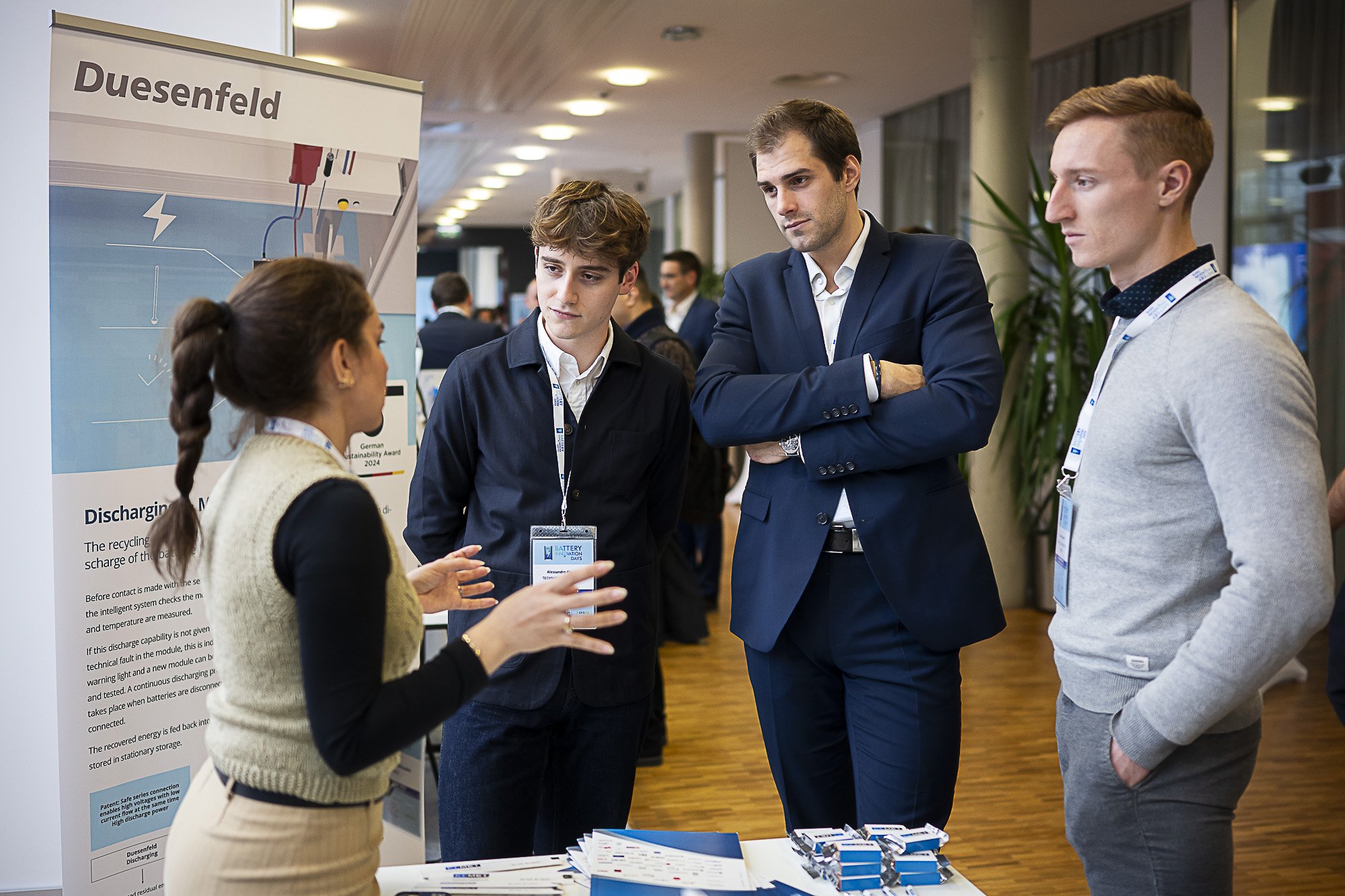 Four people engaged in a discussion at a conference, standing near a poster presentation about battery sustainability. One woman explains while three men listen attentively; they are in a spacious, well-lit conference hall.