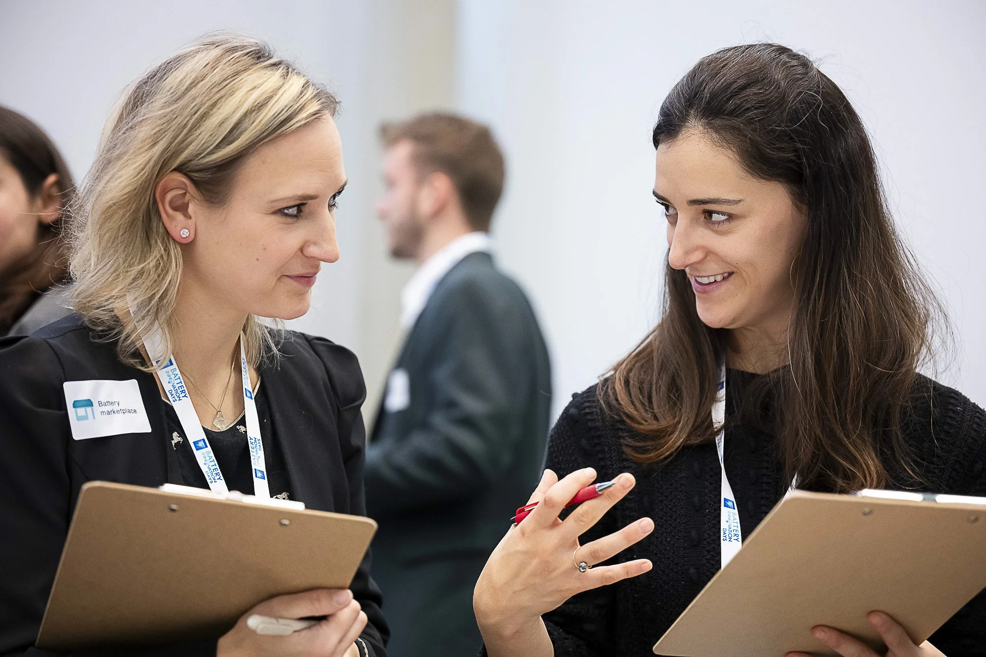 Two women wearing conference badges are engaged in conversation, pointing at a tablet. In the background, other attendees are visible.