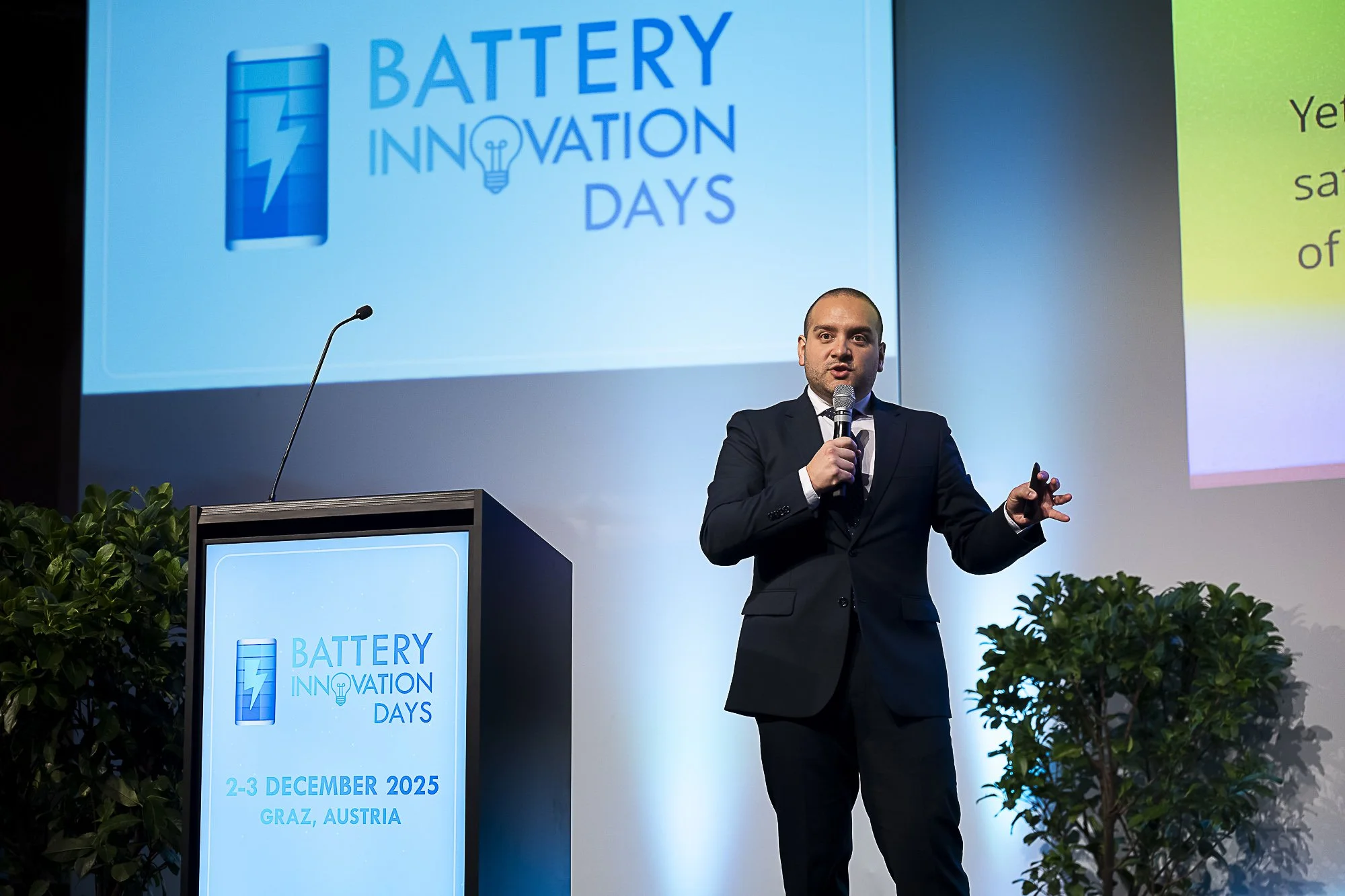A man in a dark suit and white shirt giving a presentation at a conference called Battery Innovation Days, held on December 2-3, 2025, in Graz, Austria. The backdrop displays the event logo, a blue battery icon with a lightning bolt, and a large proj