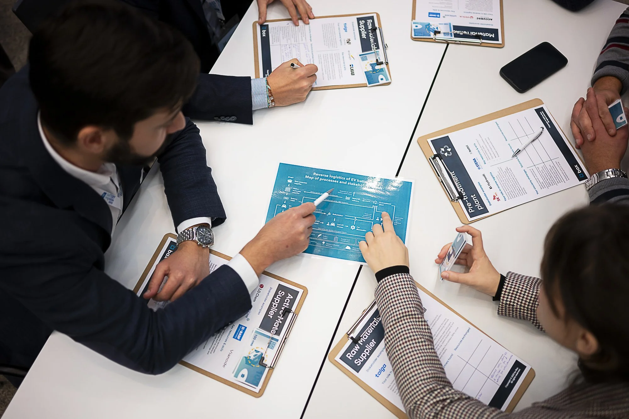 People sitting around a table at a business meeting, discussing documents and filling out forms with clipboards and pens.