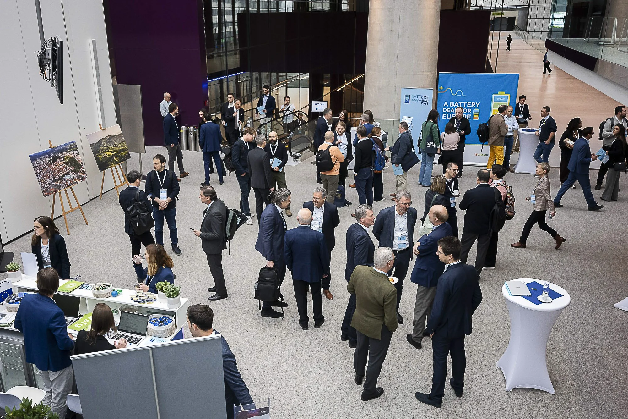 Conference attendees mingling and networking in a spacious, modern atrium with exhibition booths and artwork displayed on the walls.