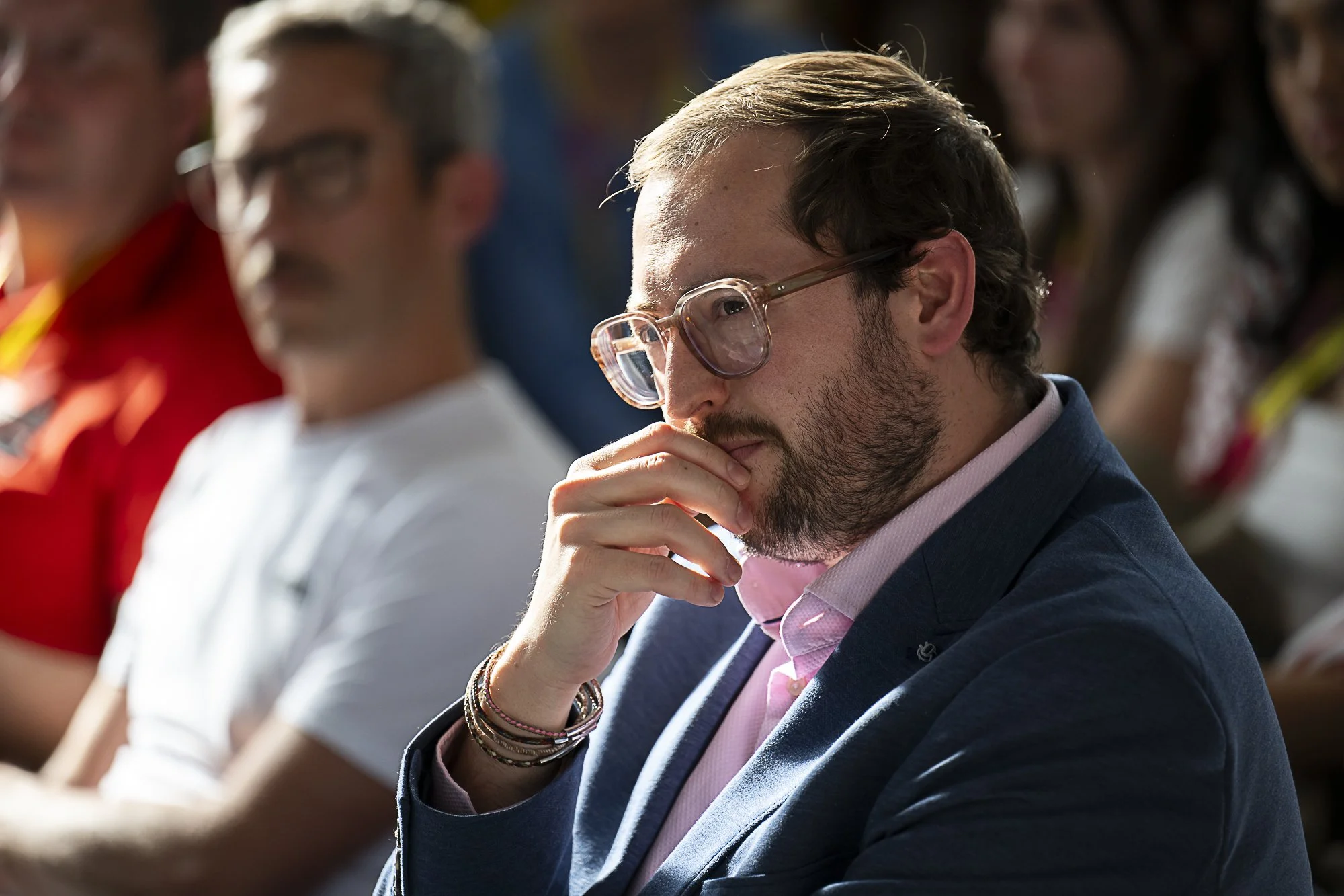 A man with glasses and a beard in a navy blue suit and pink shirt, sitting with his hand on his chin, appearing thoughtful. Several other people are seated around him, blurred in the background.