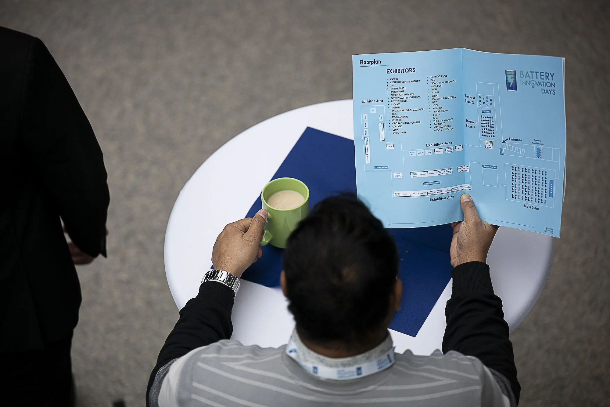 People at a round table looking at a blue floorplan for 'Battery Innovation Days' event, with one person holding a green cup.