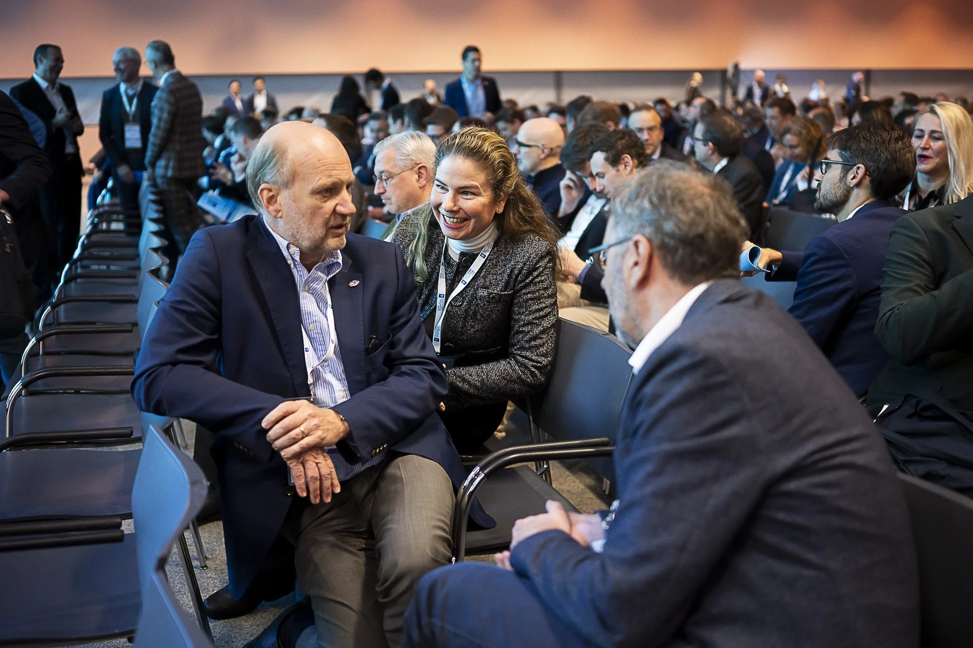 A man and a woman engaged in a conversation during a conference. The conference room is filled with attendees, most of whom are seated and talking in small groups. The background shows a large screen and more people standing and mingling.