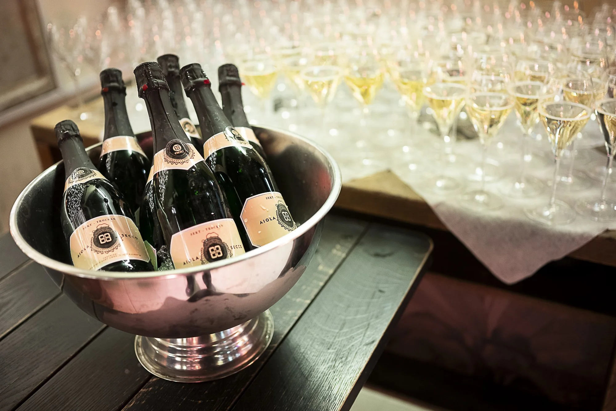 A silver bowl filled with bottles of sparkling wine or champagne chilling in ice on a dark wooden table, with glasses of champagne in the background.