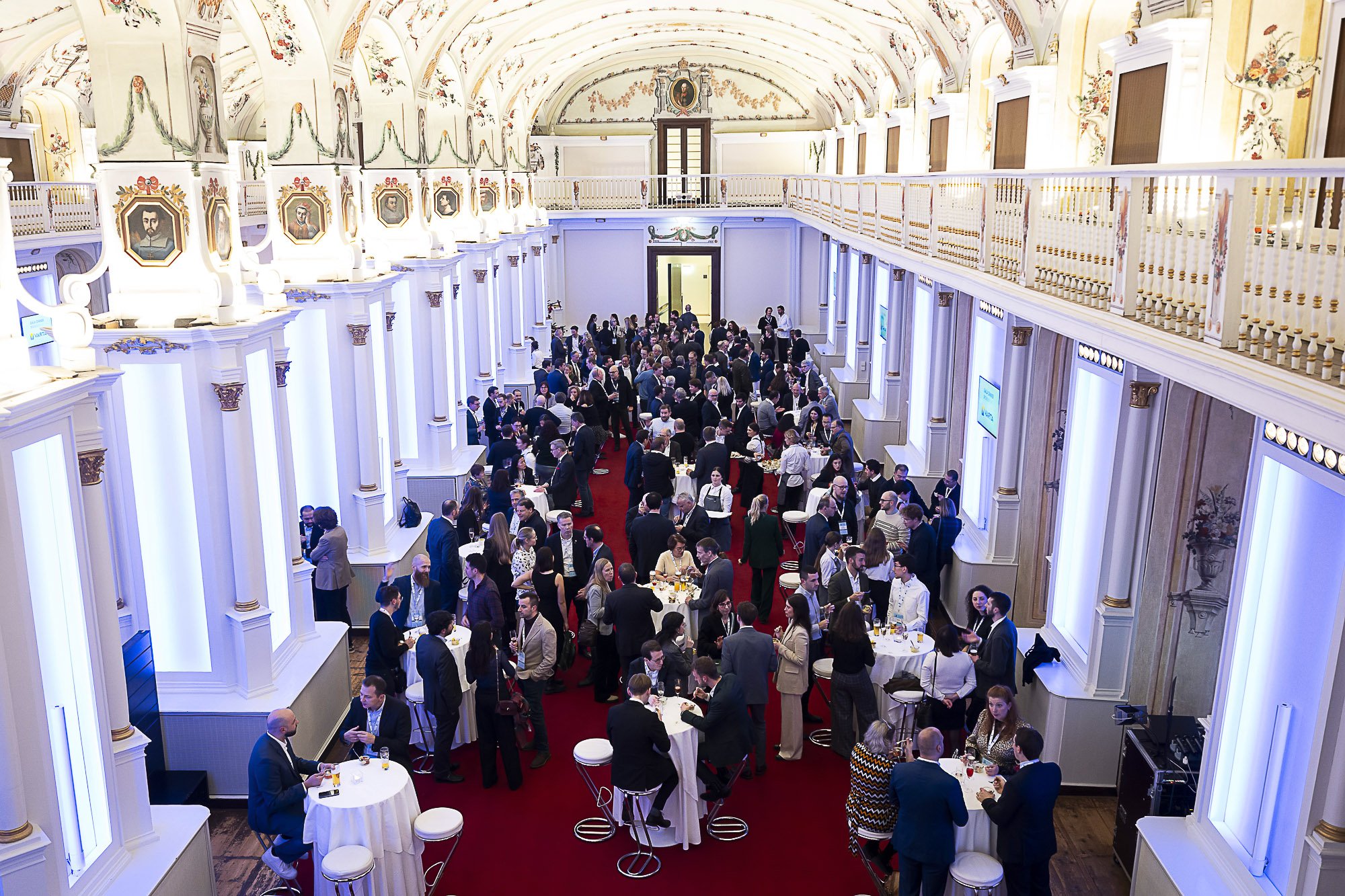 A large, ornate banquet hall filled with people attending a social event or conference. The hall has high, decorated ceilings with floral and classic mural paintings, and walls with portrait paintings framed in decorative moldings. Attendees are gath