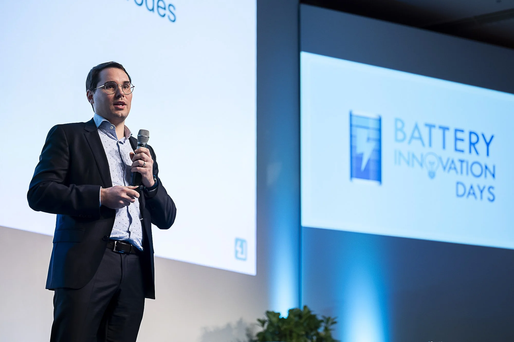 A man in a suit with glasses holding a microphone, standing on stage at a conference called Battery Innovation Days.