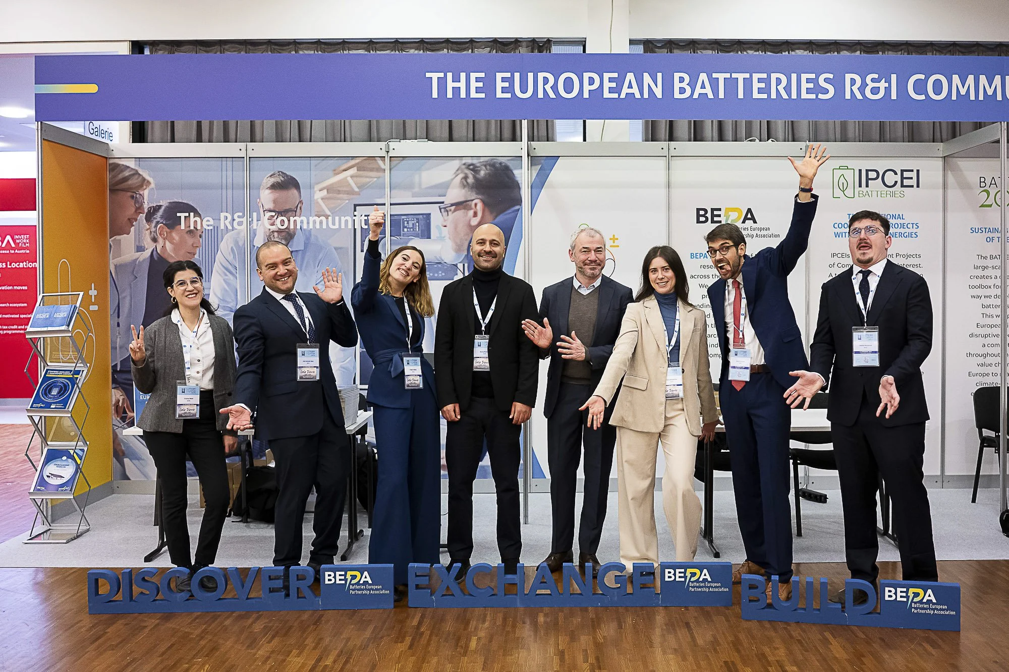 Group of seven people in business attire standing inside a booth at a conference or event, smiling and gesturing towards the camera, with a large sign above reading 'THE EUROPEAN BATTERIES R&I COMMUNITY' and a blue display with words 'DISCOVER,' 'EXC