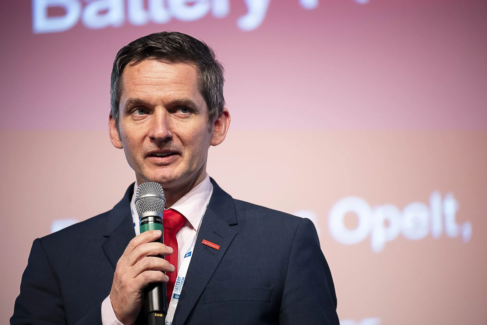A man in a suit holding a microphone speaking at an event, with a pinkish background and partial text visible in the background.
