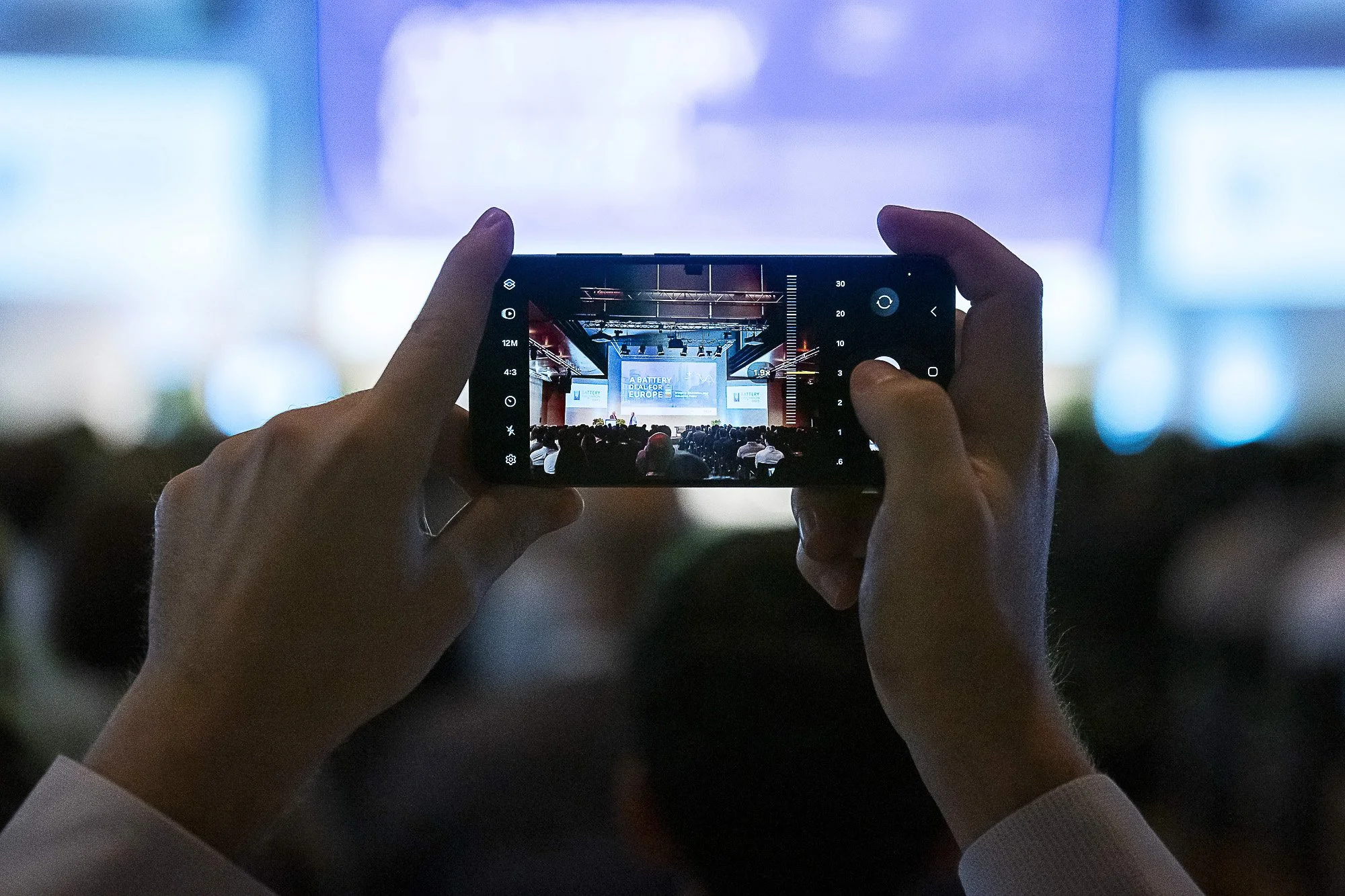 A person is holding a smartphone and taking a photo of a conference or presentation on a stage with a large audience.