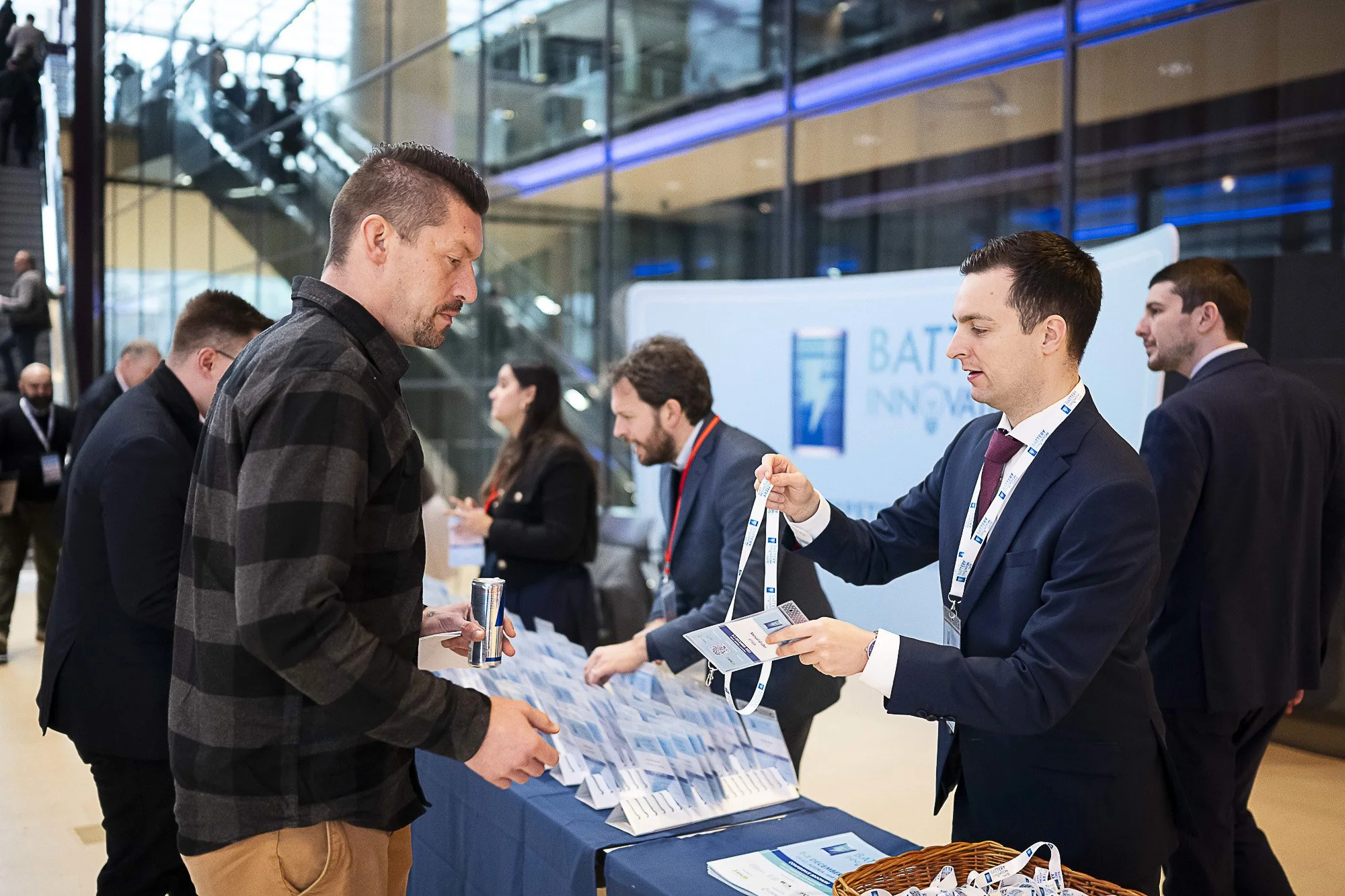 A man in a plaid shirt talks to a woman in a navy blazer at a convention or expo booth, with pamphlets and a basket on the table in front of them.