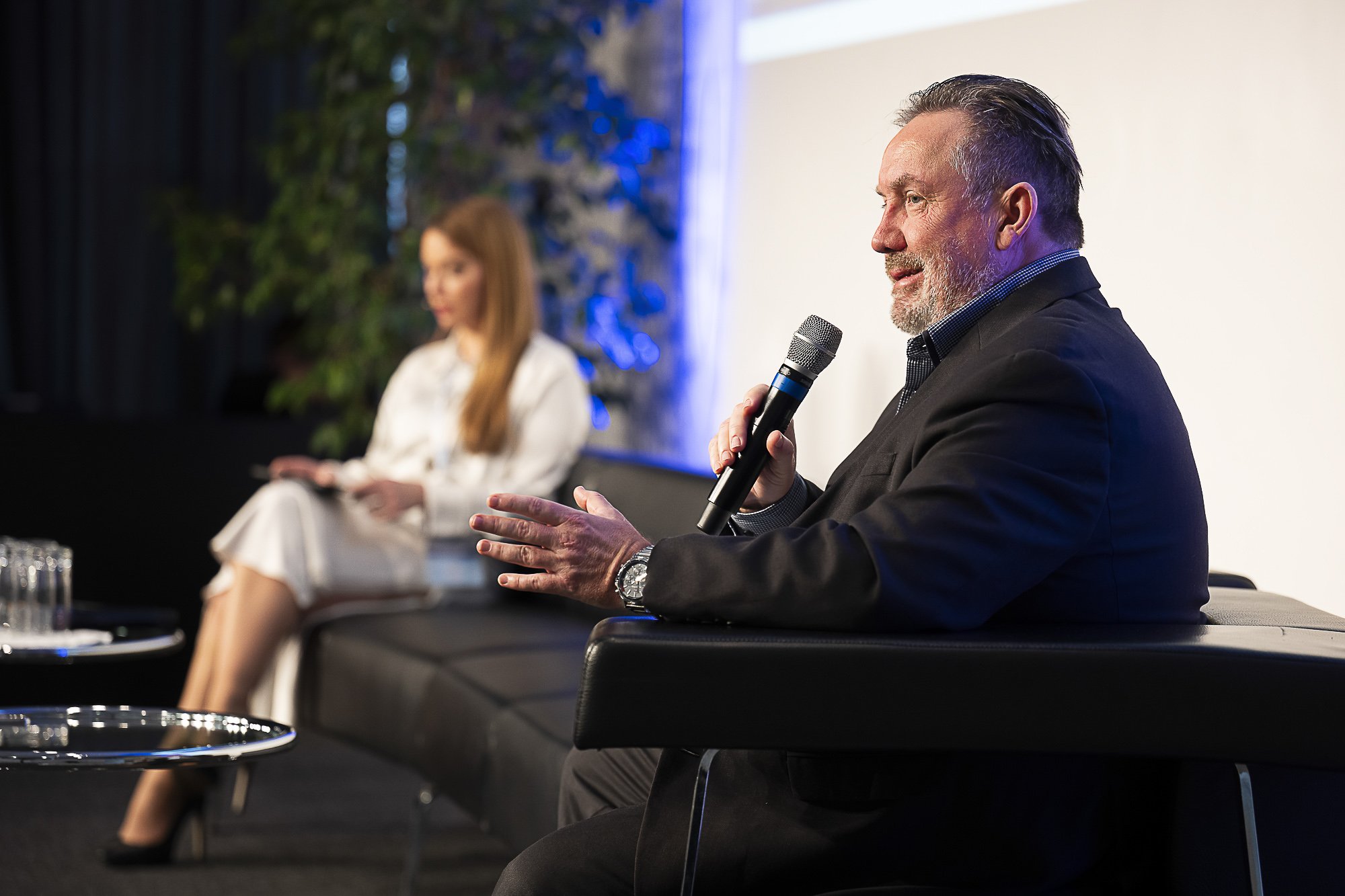 A man with a grey beard and slicked-back hair speaking into a microphone during a panel discussion. A woman in white attire is seated in the background, blurred out.