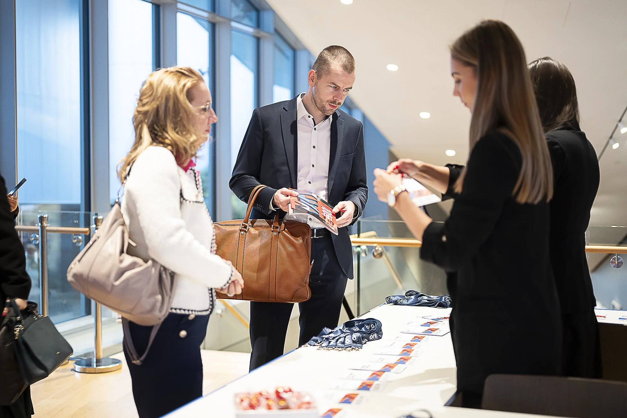 People checking in at a registration table, with event lanyards on the table, inside a modern building with large windows.