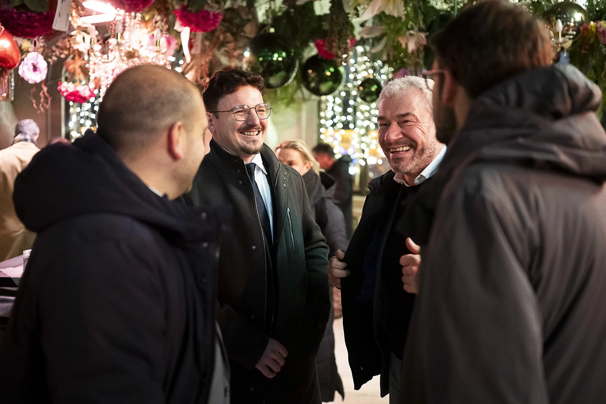 Four men are having a conversation at a festive indoor event decorated with string lights, greenery, and ornaments. All four are smiling.
