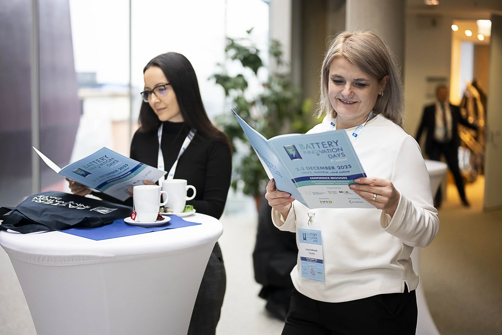 Two women at a conference stand beside a small round table reading conference brochures titled 'Battery Innovation Days' for December 2-3, 2025, in Graz, Austria.