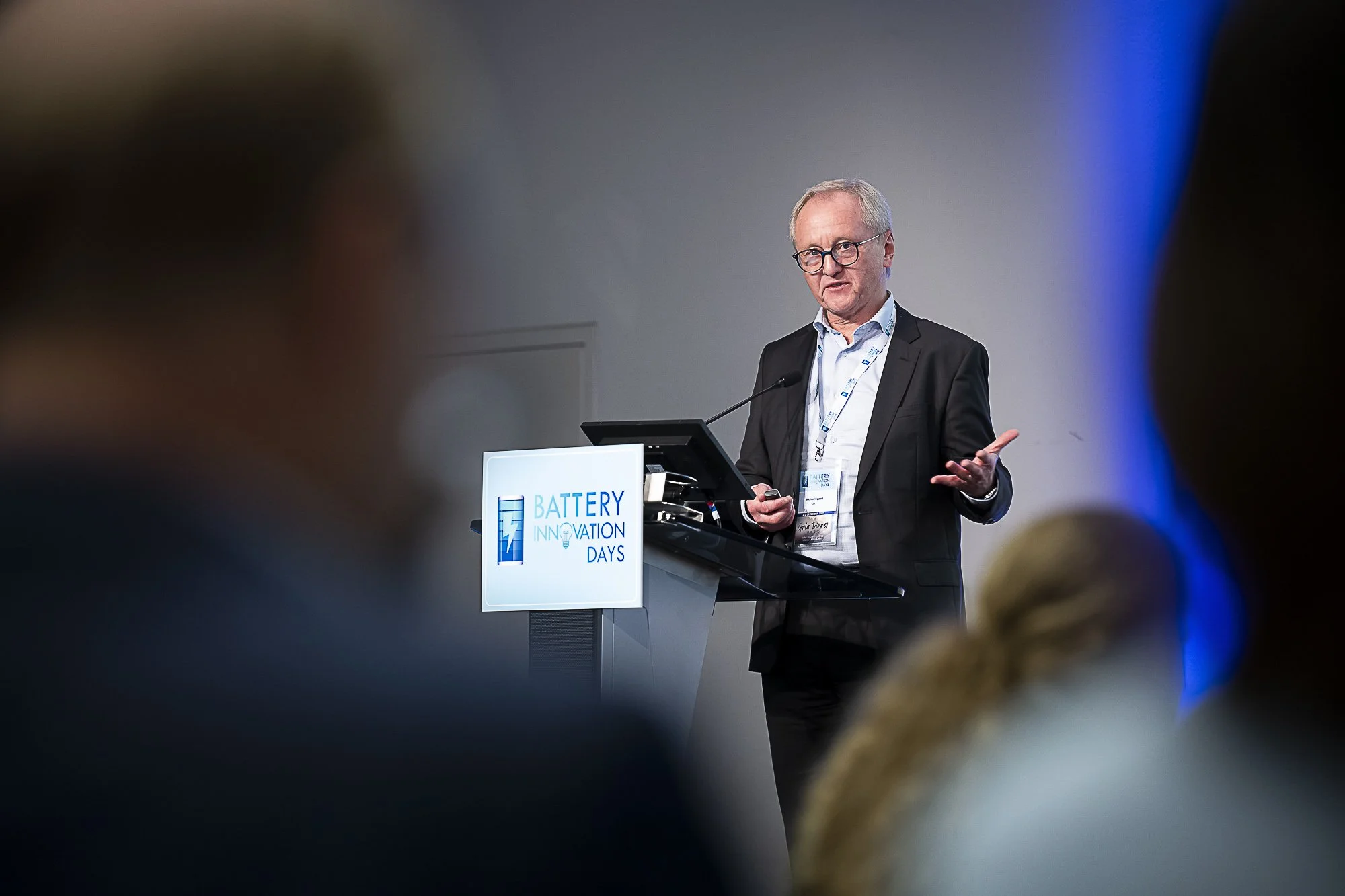 A man in a suit and glasses speaking at a podium with a sign that reads 'Battery Innovation Days.' He appears to be giving a presentation or speech, gesturing with his right hand.