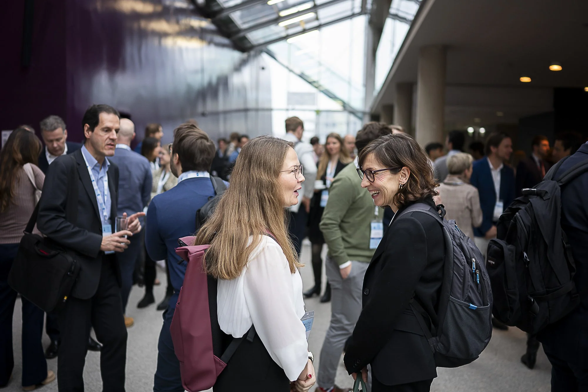 A group of people at a conference or networking event, engaged in conversations in a modern indoor space with large windows and high ceilings.