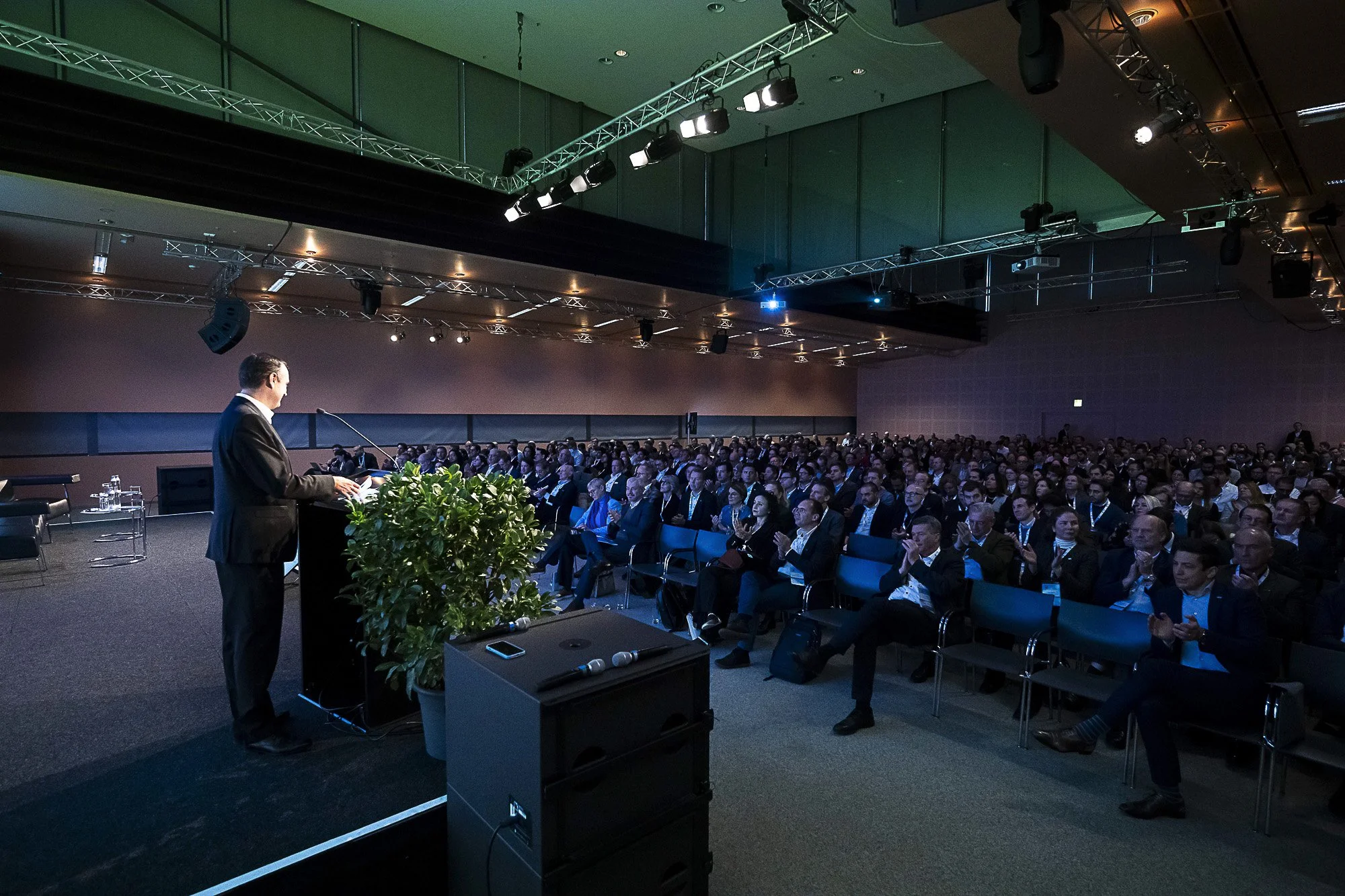 A man in a suit delivering a speech at a conference or seminar, with a large audience seated and clapping. The event is held in a dimly lit auditorium with stage lighting and equipment overhead.