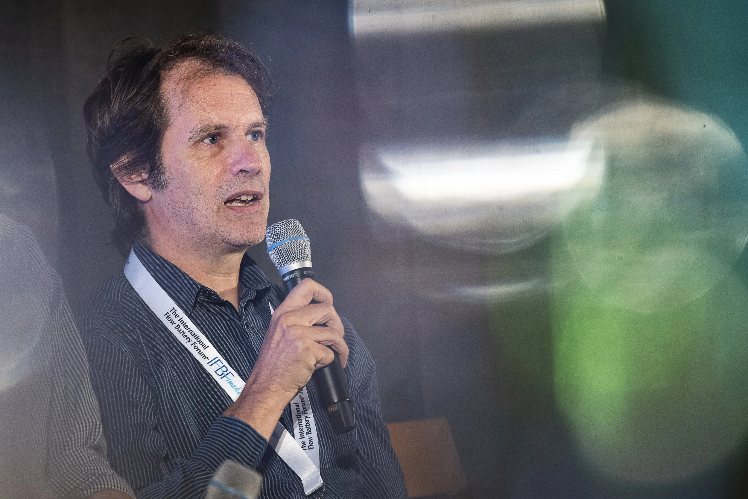 A man with brown hair holding a microphone, wearing a black and white striped shirt and a lanyard, speaking at an event with a blurred background and colorful bokeh lights.