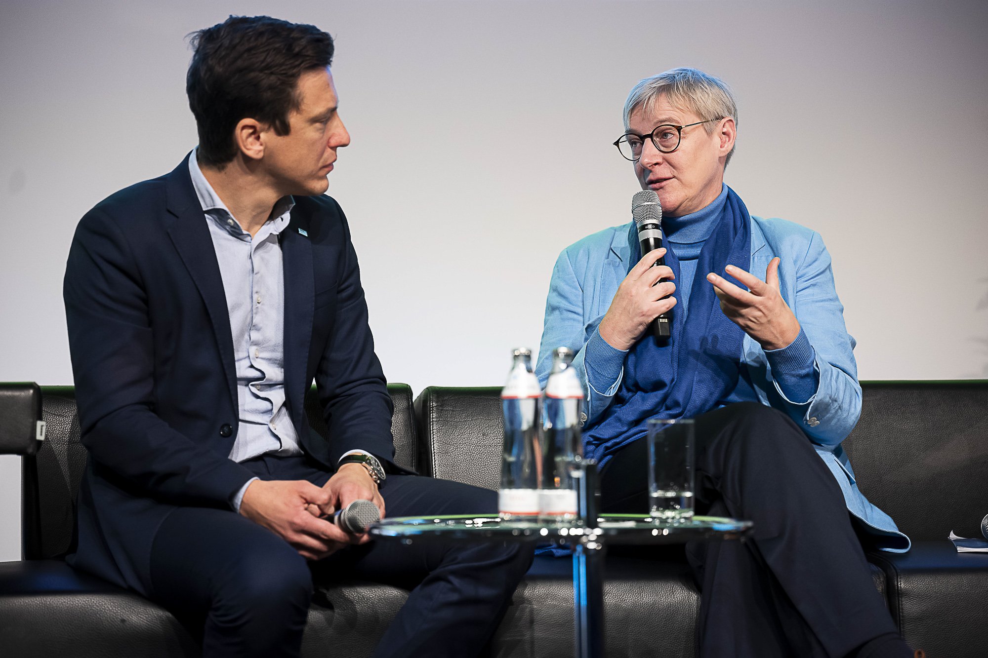 Two men sitting on a panel discussion, one holding a microphone and speaking, the other listening attentively. They are dressed in business attire and are seated on a black leather couch with water bottles and microphones on a table in front of them.