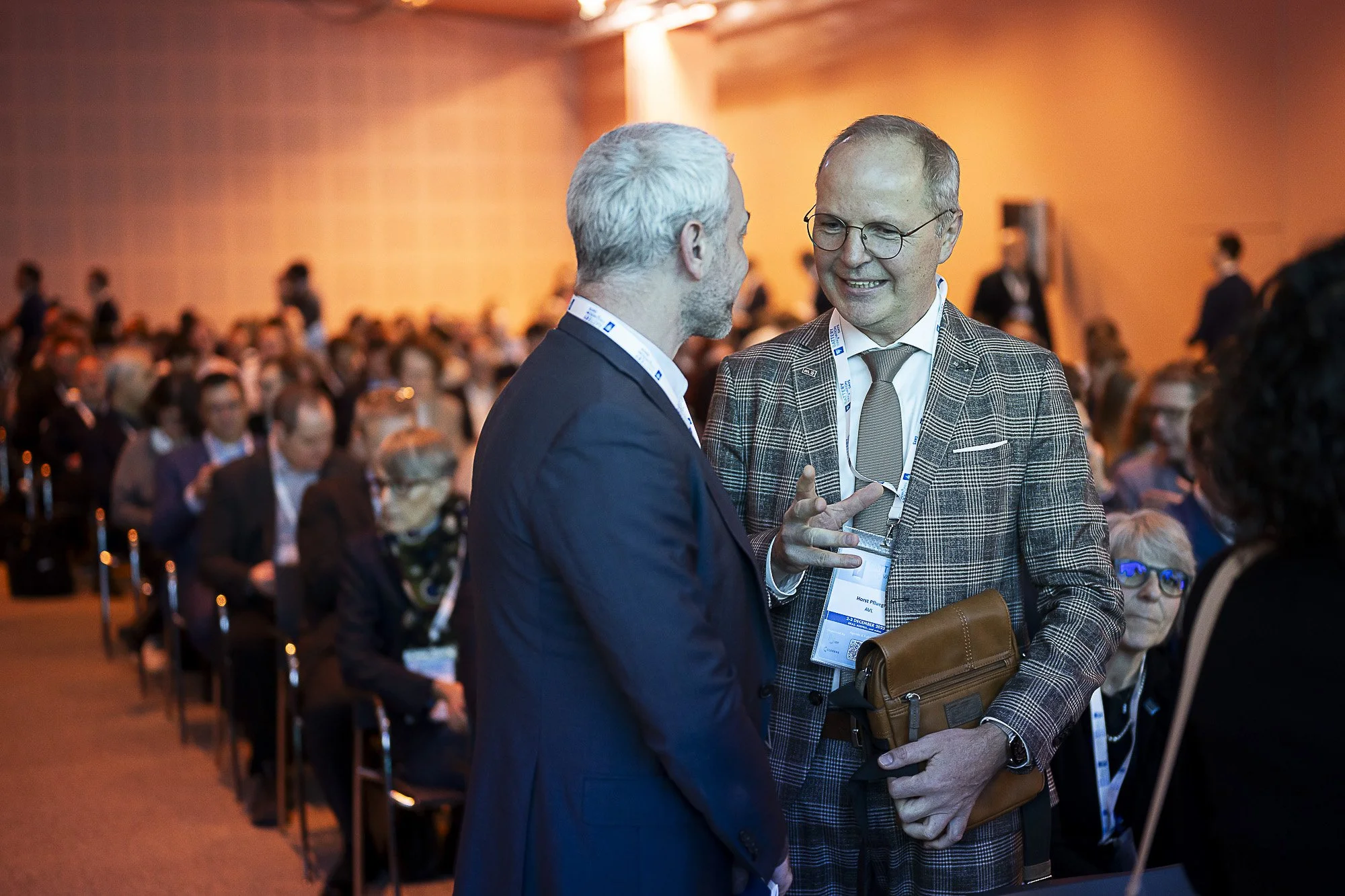Two men are engaged in a conversation at a conference with many seated attendees in the background. One man has silver hair and a beard, wearing a dark suit, while the other has glasses, a checkered jacket, and is holding a brown bag.