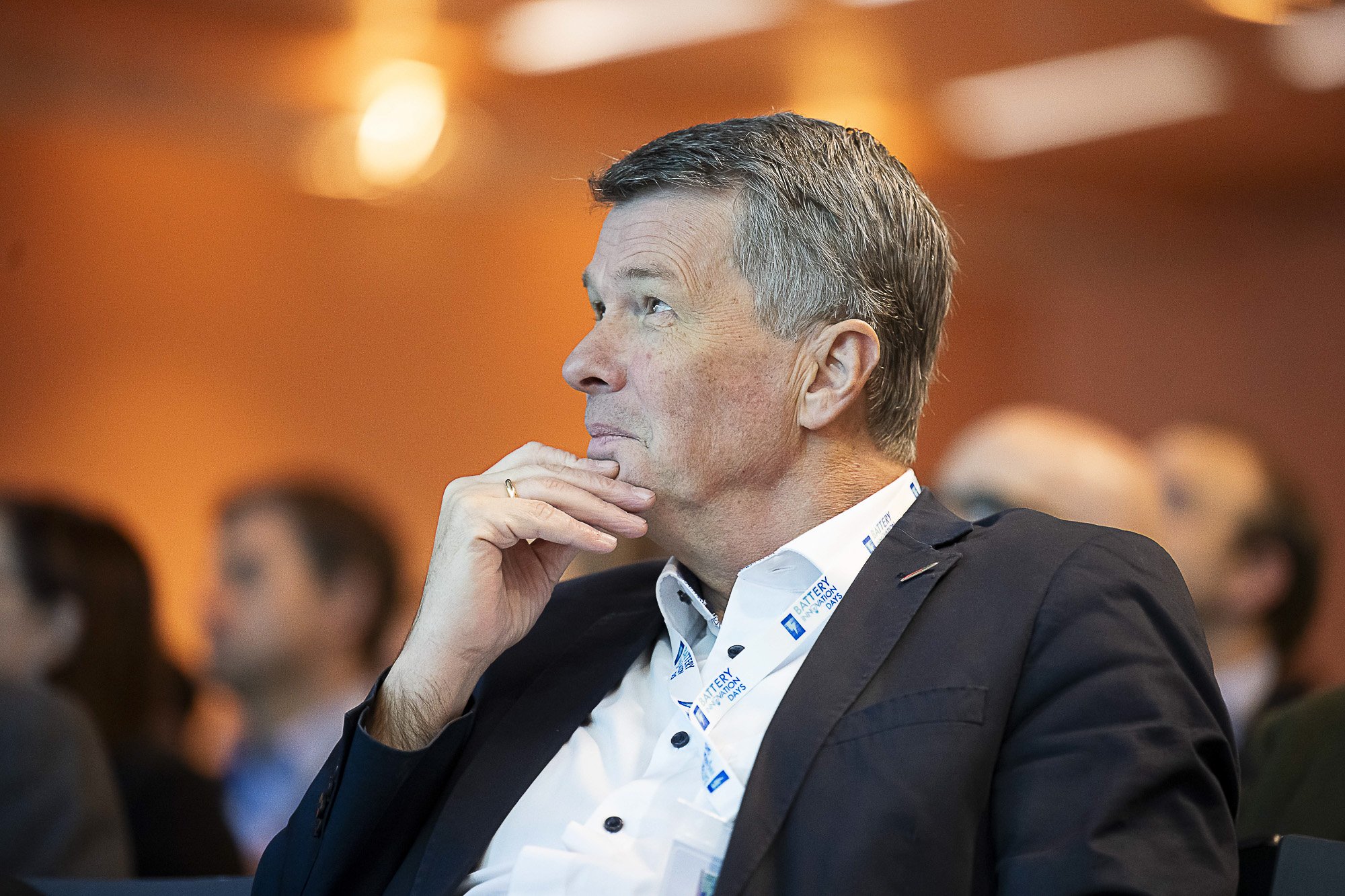 A man in a suit with gray hair in profile, seated at an indoor event, resting his chin on his hand, with a thoughtful expression. He is wearing a lanyard with text that includes 'Battery' and 'Days' around his neck. The background is blurred with war