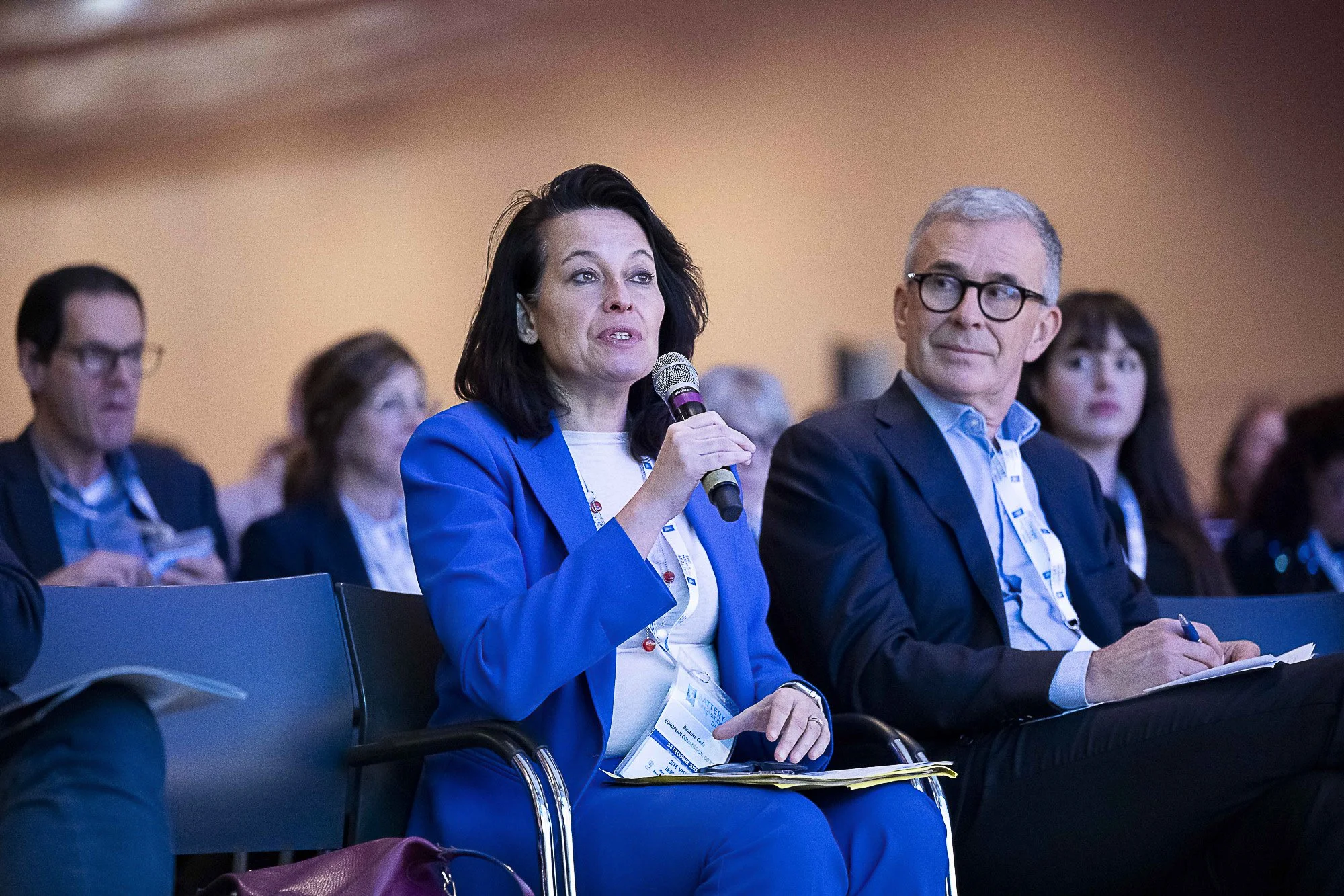 A woman in a blue blazer speaking into a microphone at a conference, with a man with glasses sitting next to her and audience members in the background.