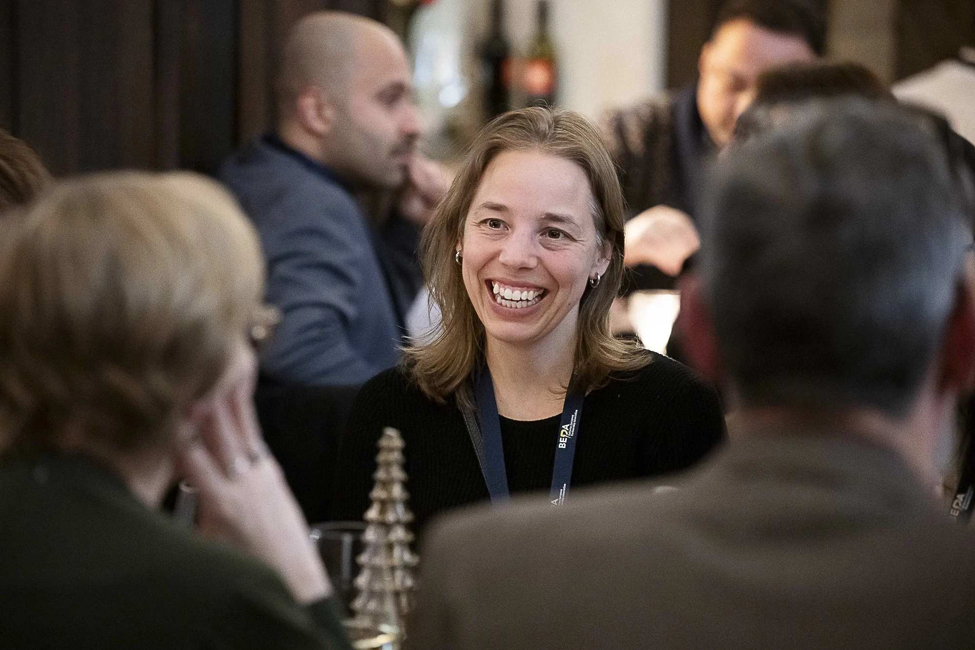 A woman with shoulder-length brown hair, smiling and wearing a black top with a blue lanyard, engaged in conversation at a conference or meeting surrounded by other people.