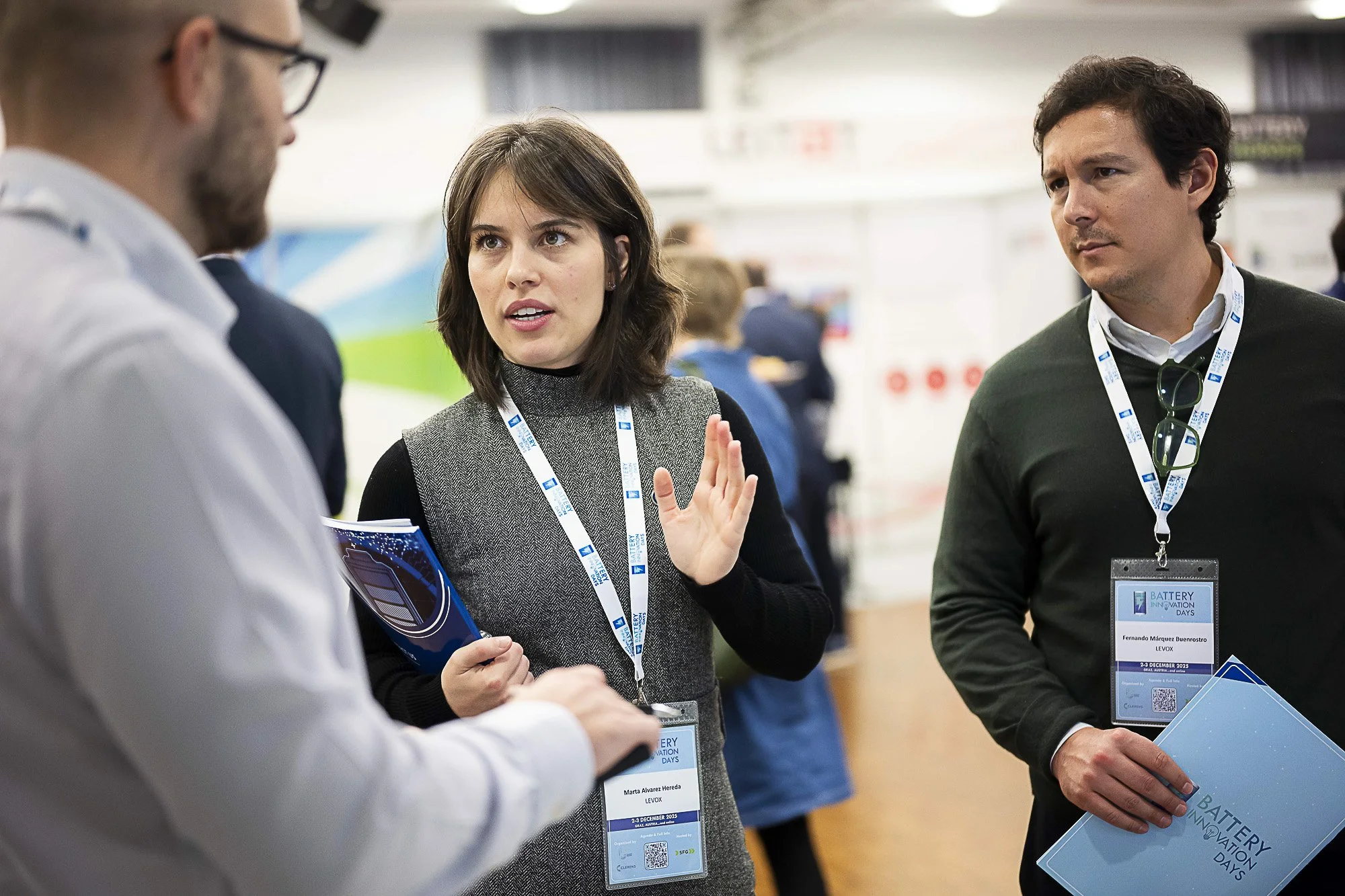 A woman with shoulder-length brown hair is speaking to two men at a conference or event. She is holding a folder or booklet and is wearing a lanyard with a badge that reads 'Battery Innovation Days' with her name 'Marta Alvarez Hereda'. The men are l