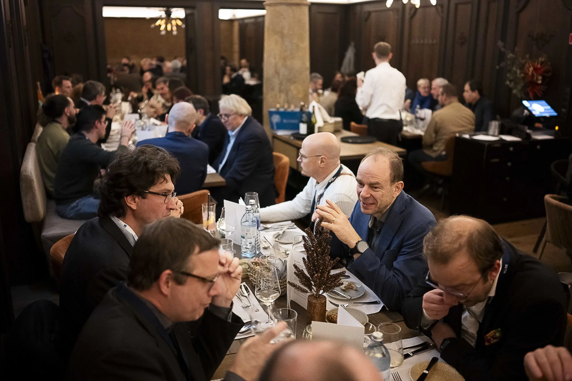 A large group of people sitting at long tables in a dining room during a formal event or celebration, with covered dishes, glasses, and centerpieces on the tables. The room has warm lighting and dark wood-paneled walls.