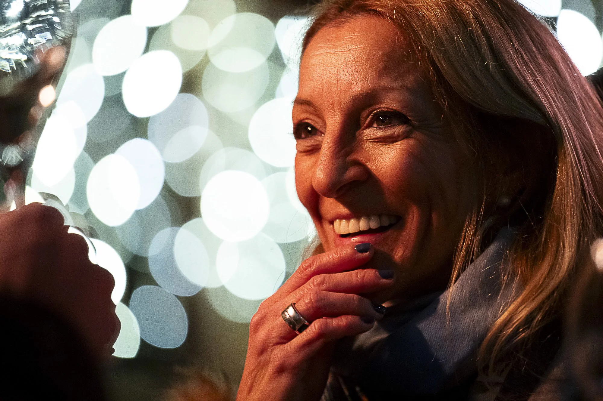 A woman with reddish-brown hair smiling and looking at her phone, with a blurred background of bright circular lights.