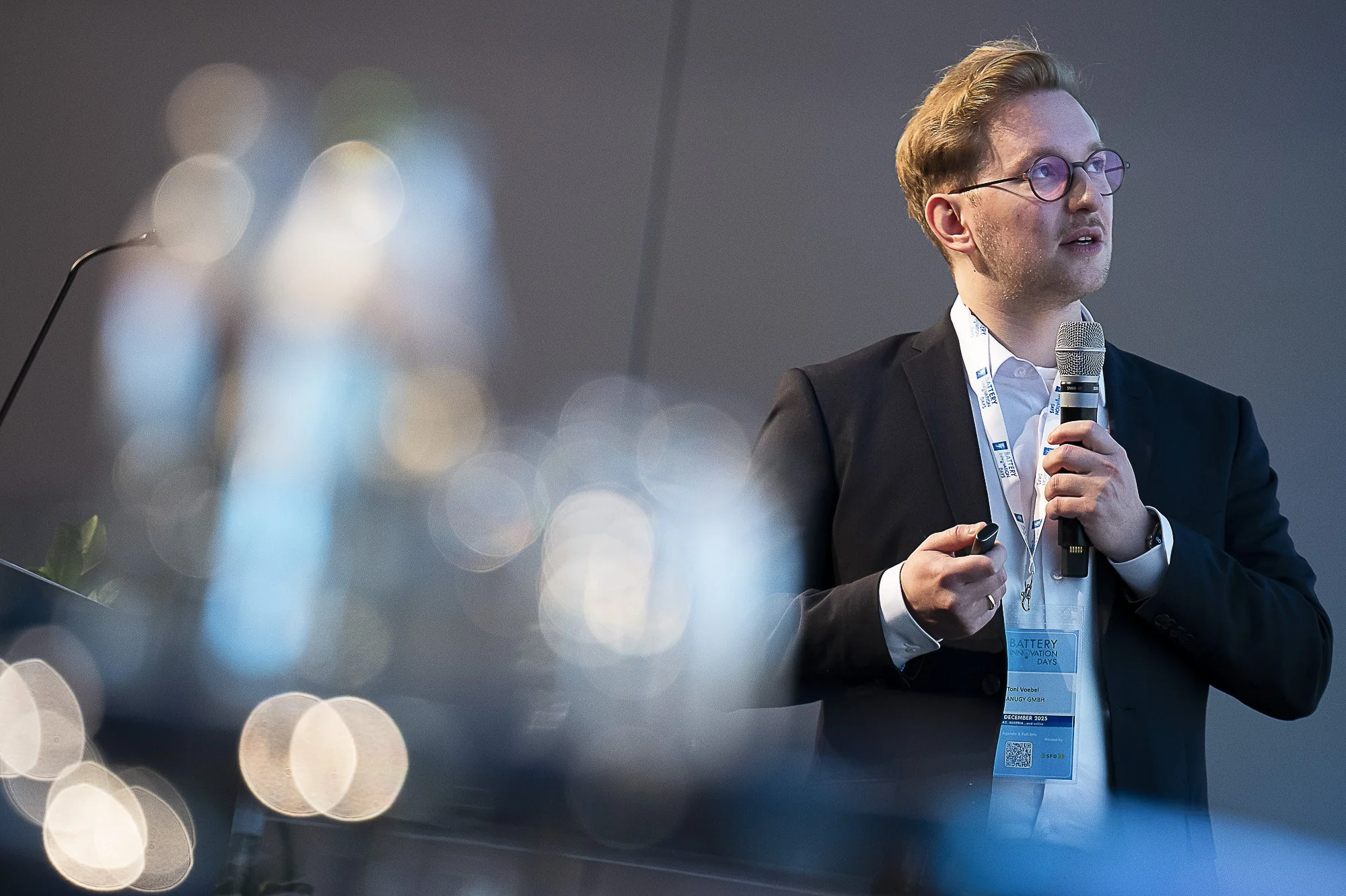 A man with glasses, dressed in a suit, holding a microphone and a remote control, giving a presentation at a conference. The background shows a blurred conference badge and conference branding.