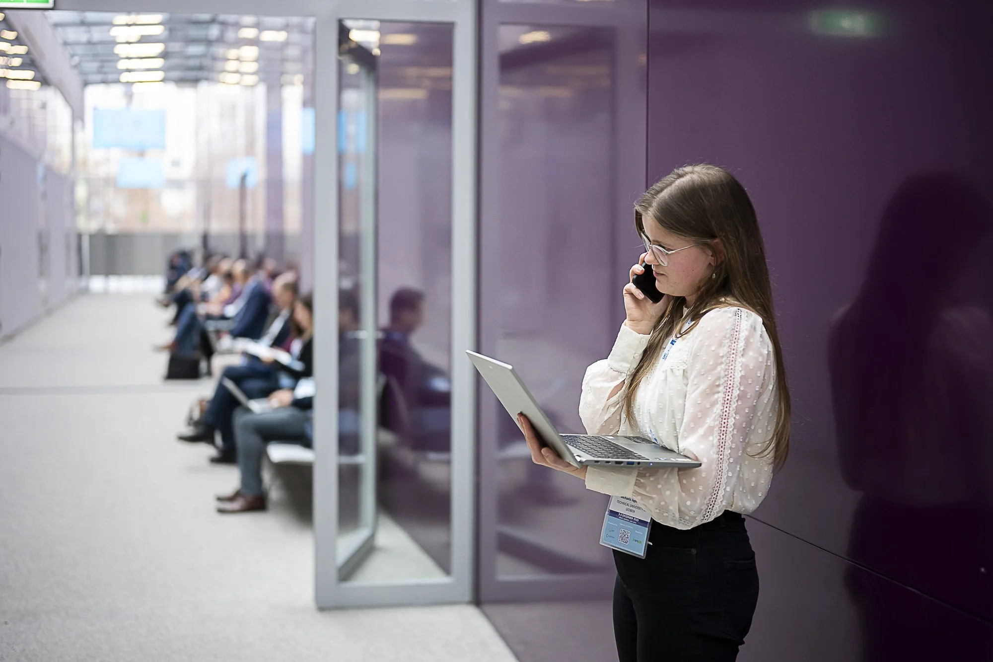 A woman holding a laptop and on the phone, standing in a modern waiting area with rows of seated people using electronic devices, purple wall, glass ceiling.