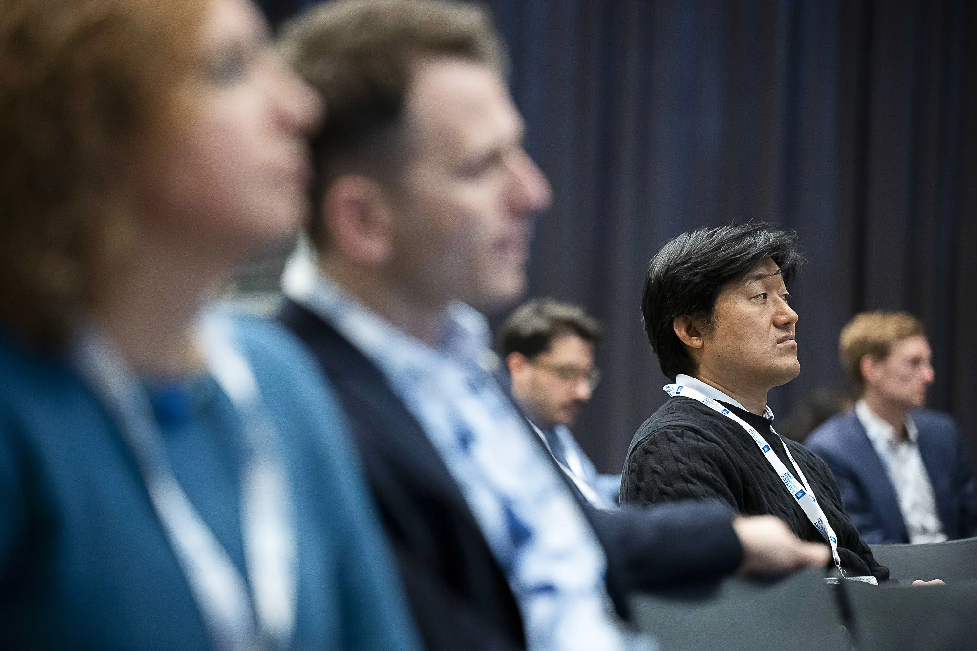 A group of people, mostly men and women, sitting attentively at a conference or seminar, dressed in business or casual attire, with identification badges around their necks, sitting in a row with a dark curtain in the background.