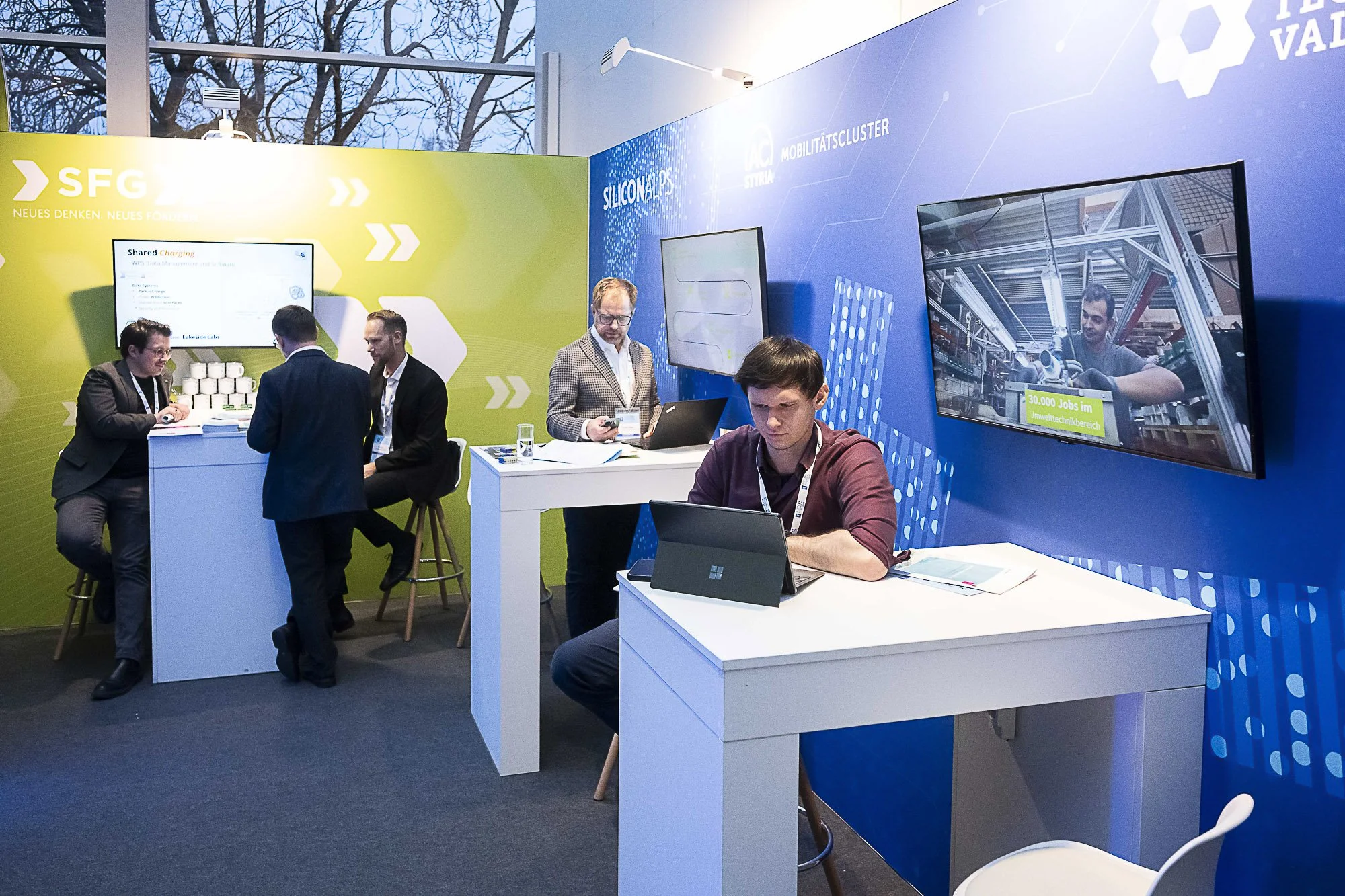 People working at a technology booth with laptops and screens in a conference setting, featuring green and blue themed walls with various digital graphics.