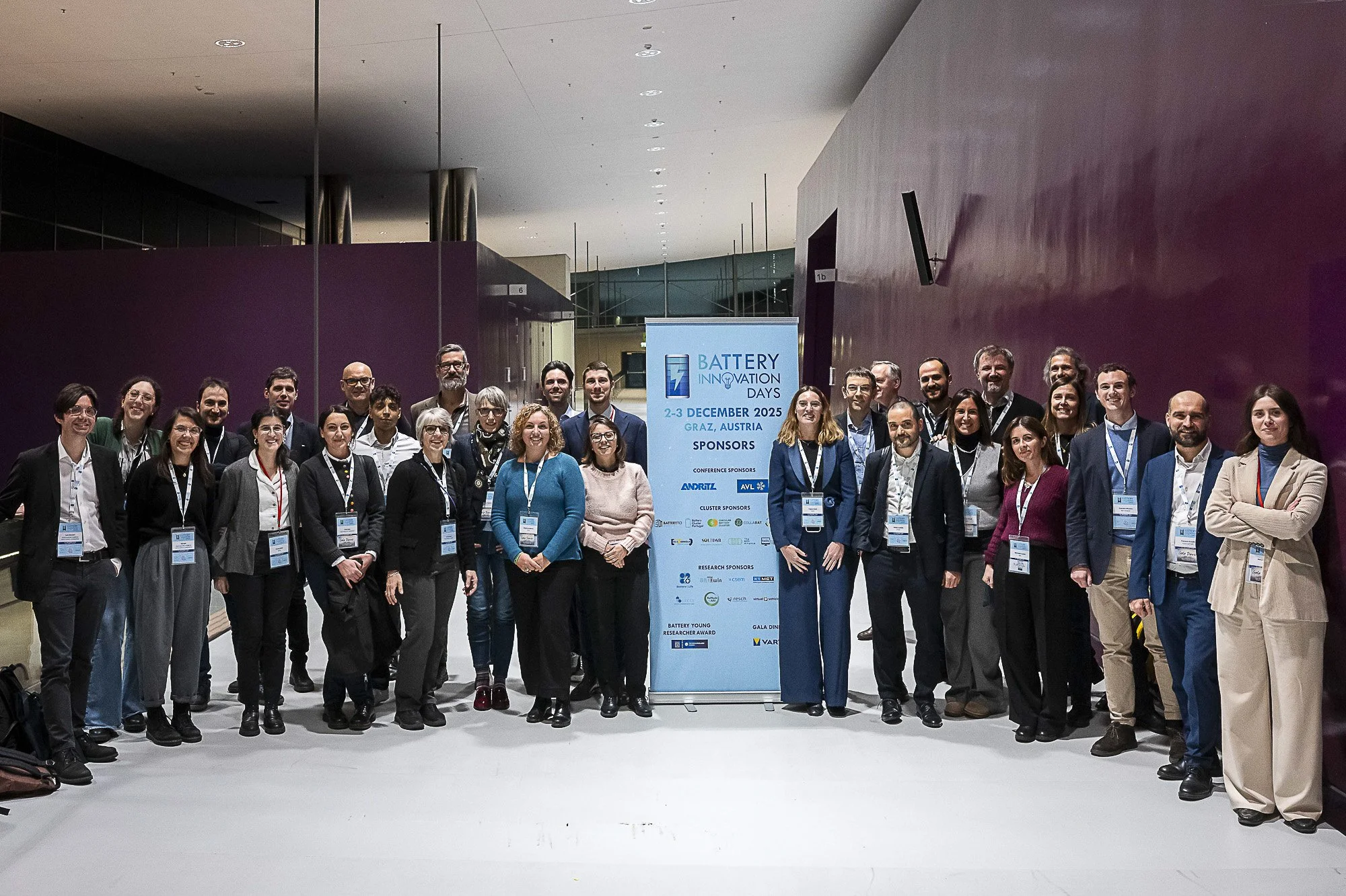 Group of people attending a conference or event, standing together in front of a sign that reads 'Battery Innovation Days, December 2-3, 2025, Graz, Austria.' The group includes men and women of various ages, dressed in business or business casual at