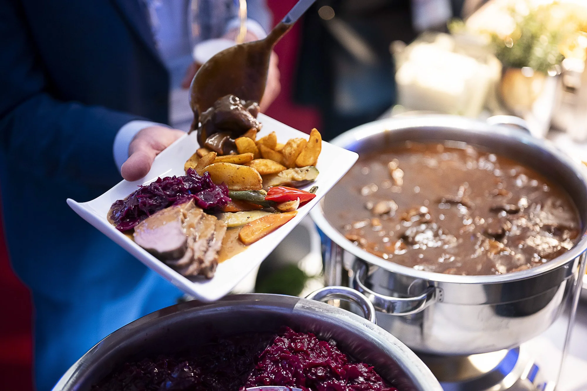 A person serving food at a buffet. They are holding a white rectangular plate with assorted dishes, including roasted vegetables, meat, and red pickled cabbage. There is a large pot of stew or soup nearby.