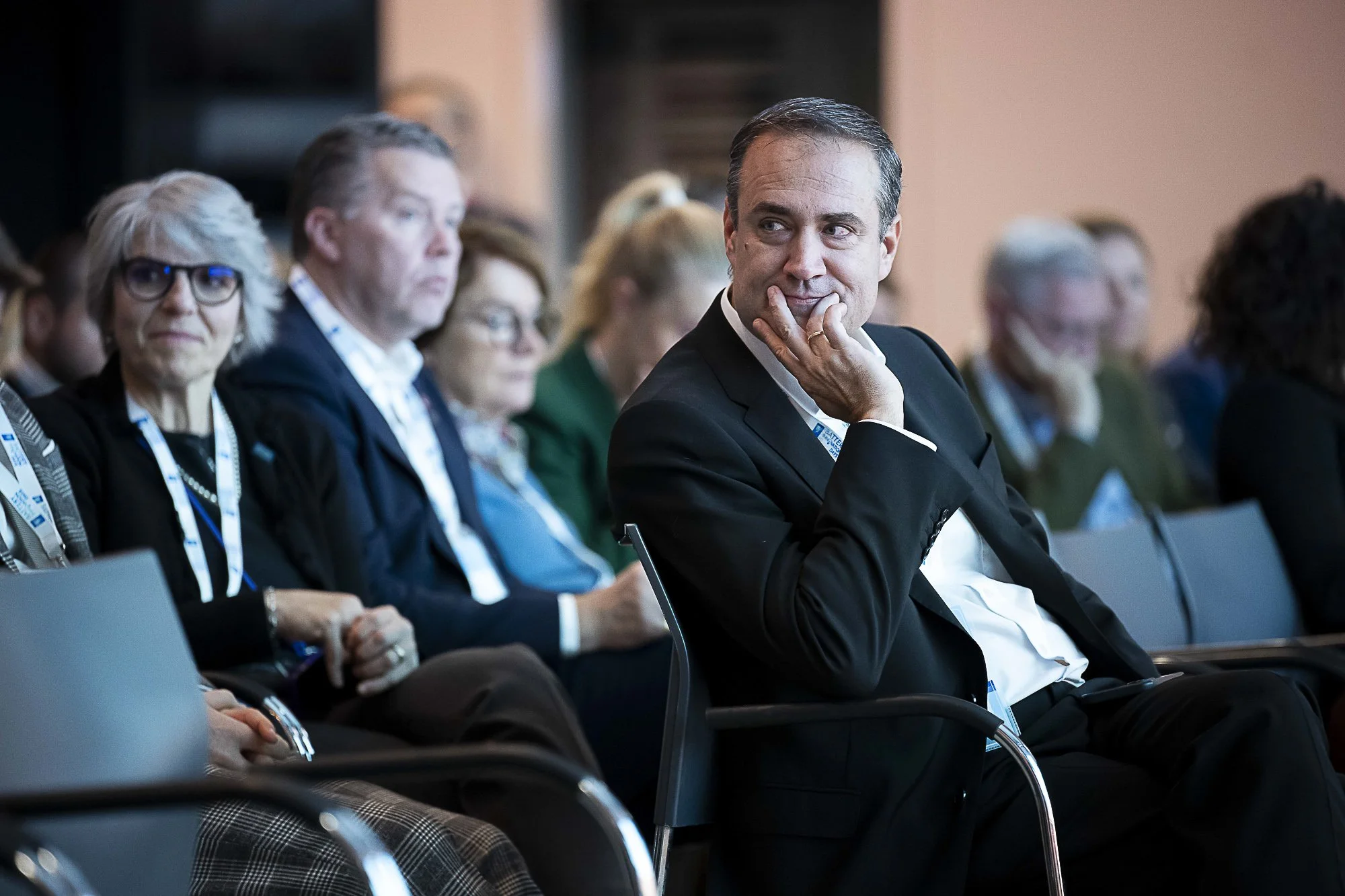 A group of people attending a conference or seminar, seated and listening. The focus is on a man in a black suit with a white shirt, sitting with his hand on his chin, looking to his right with a thoughtful expression.