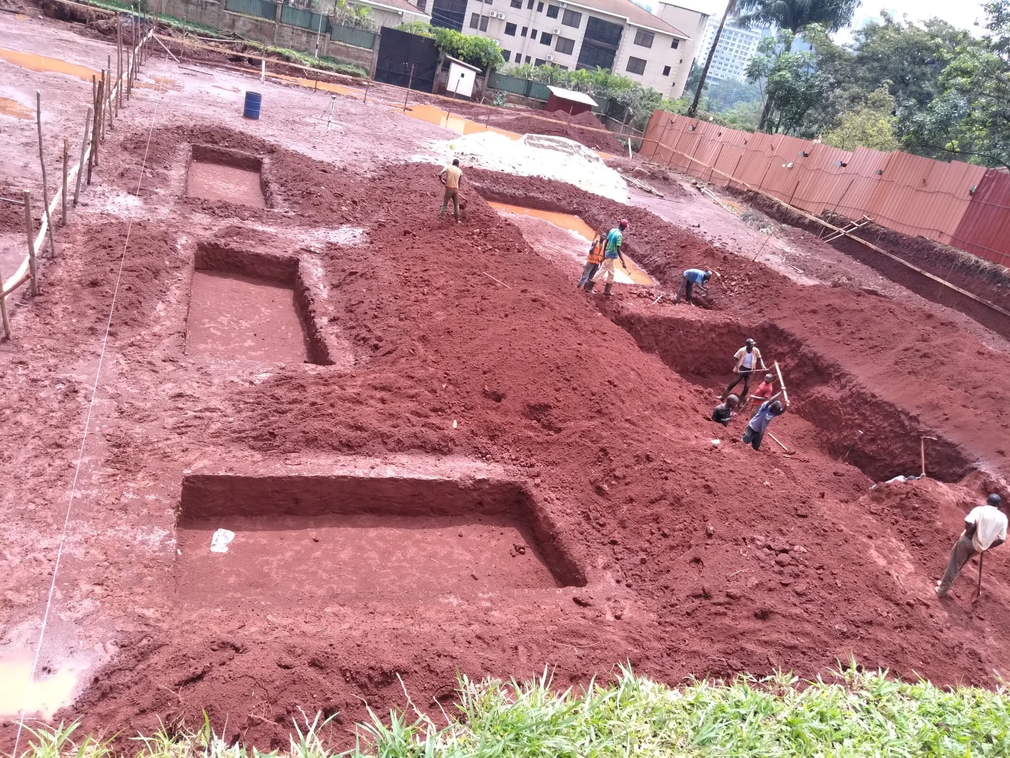 Construction workers digging and excavating on a muddy construction site with red soil, in an urban area with tall buildings and trees in the background.