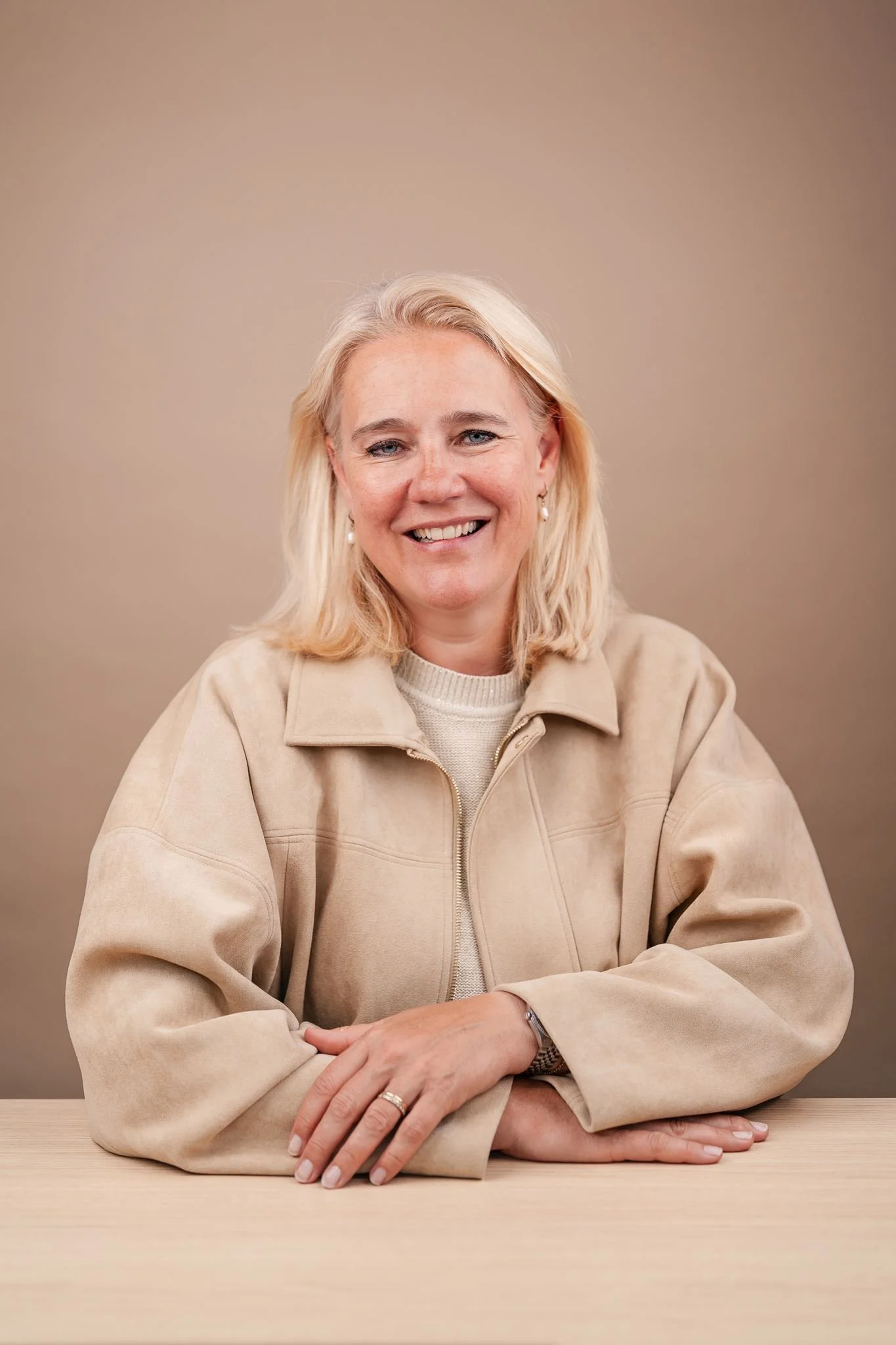 Een lachende vrouw met blond haar, gekleed in een beige jas en wit trui, gezeten achter een lichtkleurige tafel met haar armen gevouwen.