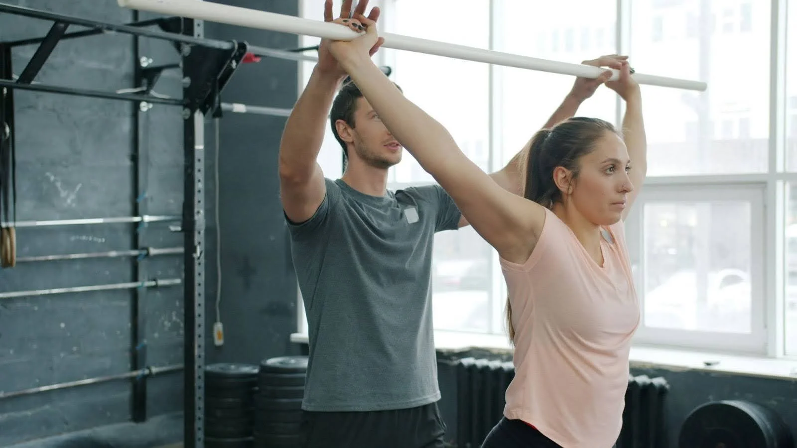 Trainer assisting a client with an overhead mobility stretch using a bar.