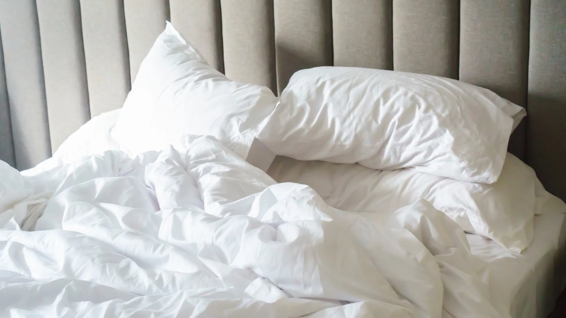 A bed with white pillows and rumpled white sheets against a padded grey headboard.