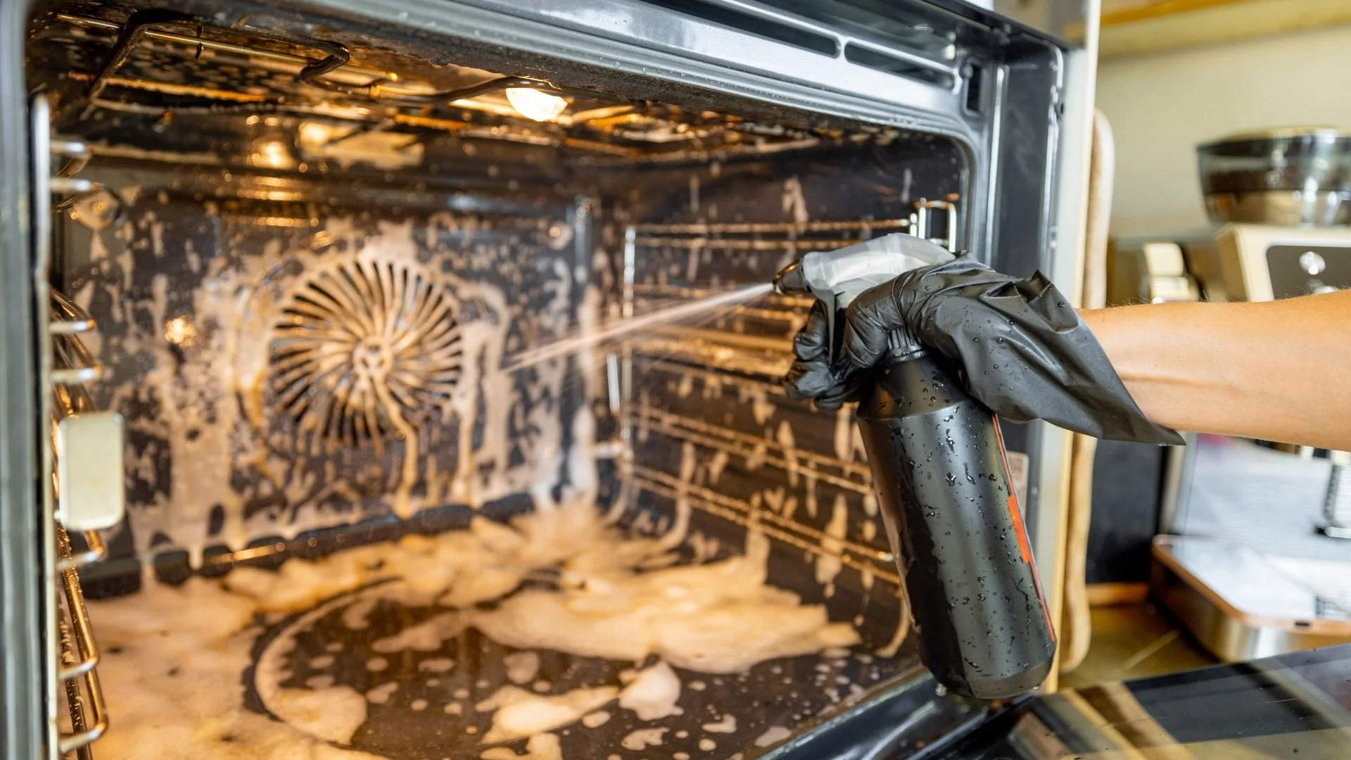 A person wearing a black glove is steam cleaning the inside of an oven with a spray bottle.