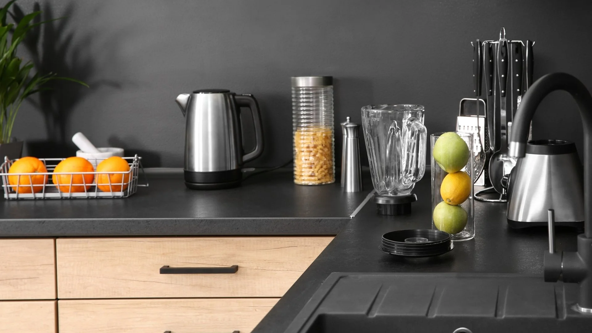 Kitchen counter with a wire basket of oranges, a stainless steel electric kettle, a container of pasta, a glass pitcher, a blender with apples and lemon, and various kitchen utensils on a dark wall background.