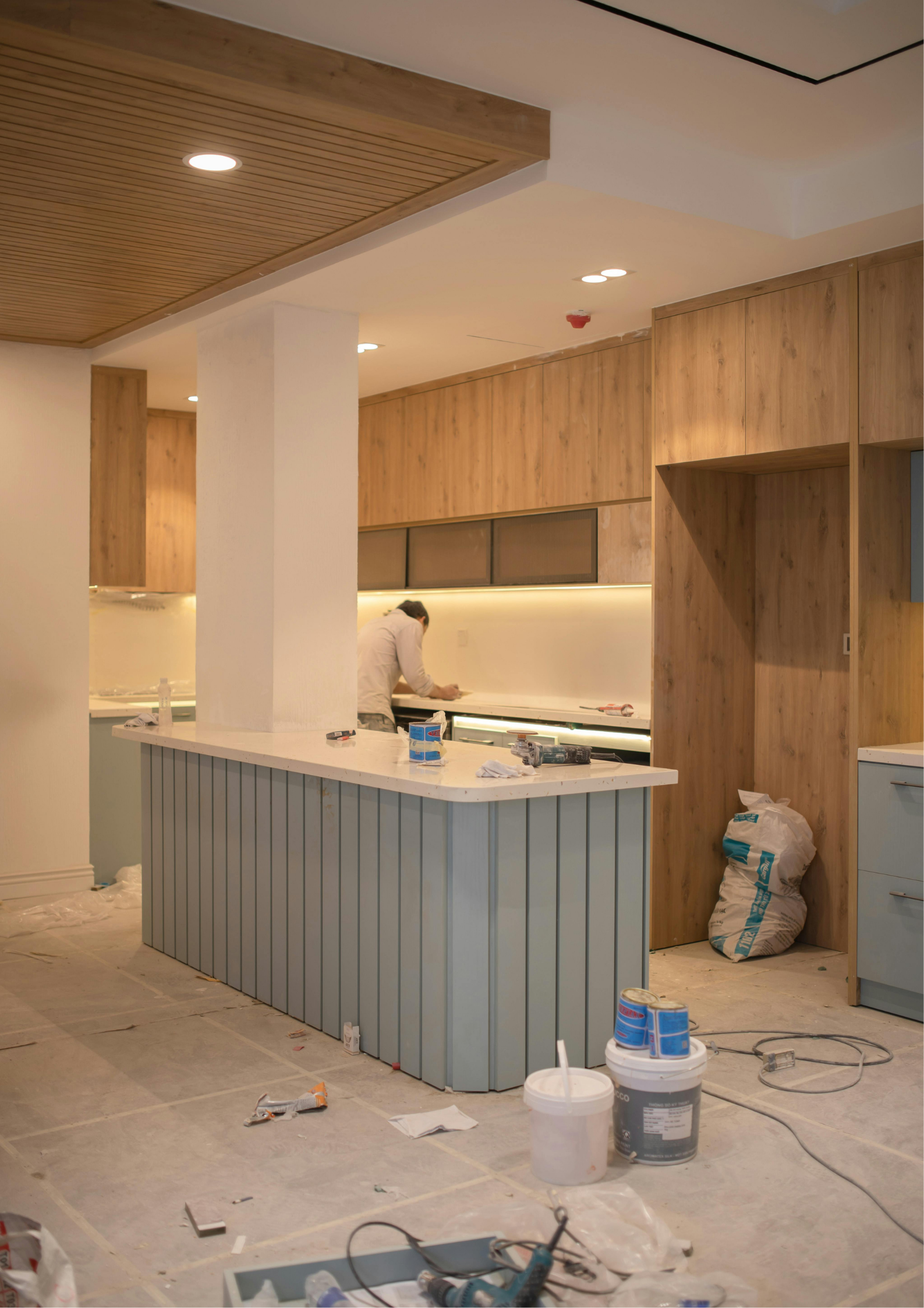 A room under renovation with a kitchen island or counter, construction tools, materials, and a person working in the background.